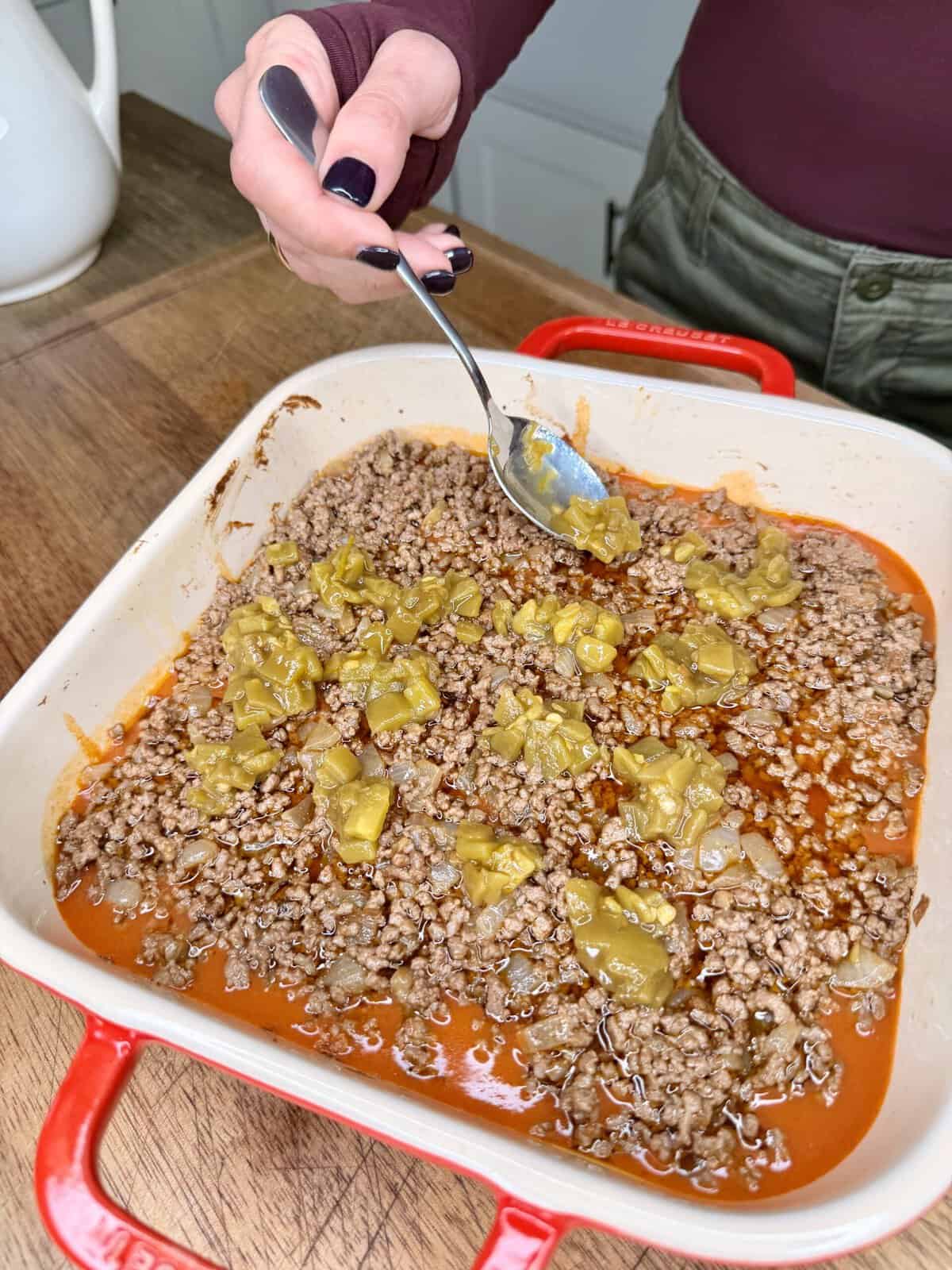 A hand holds a spoon over a red baking dish filled with cooked ground beef, chopped green chiles, and sauce on a wooden surface.
