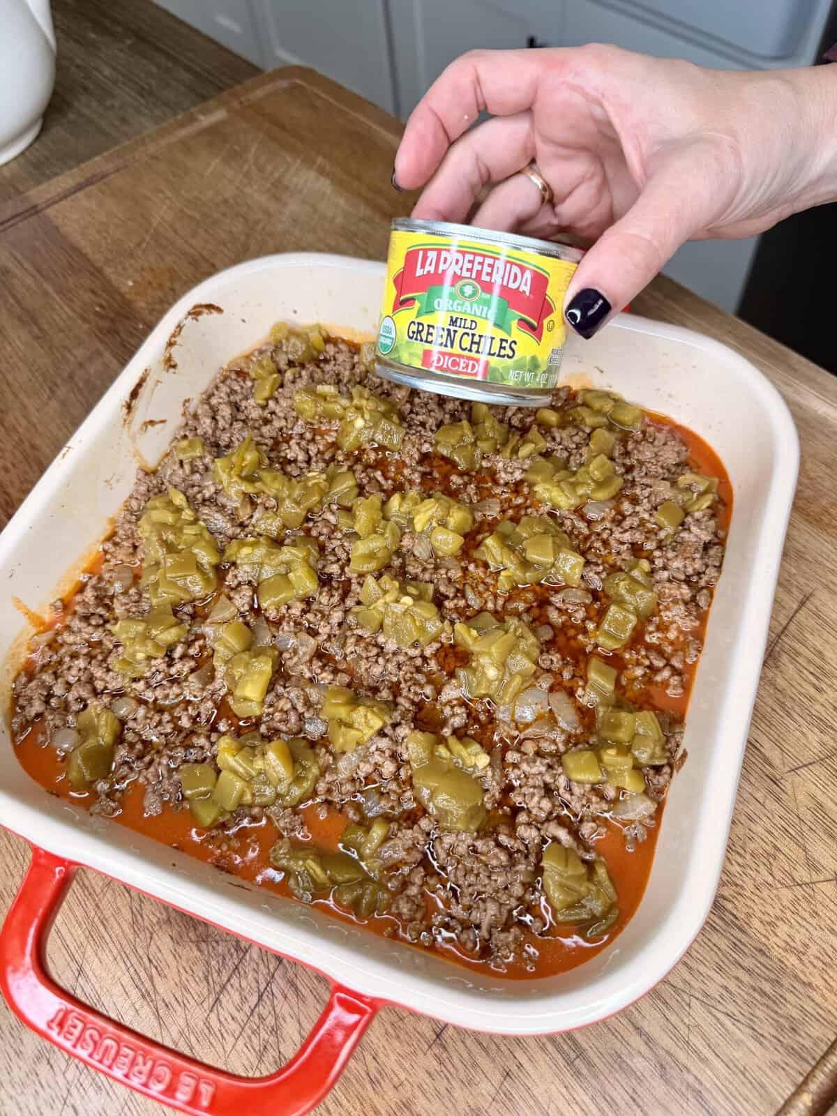 A hand holds a can of La Preferida green chiles over a baking dish filled with cooked ground beef and green chiles. The dish sits on a wooden countertop.