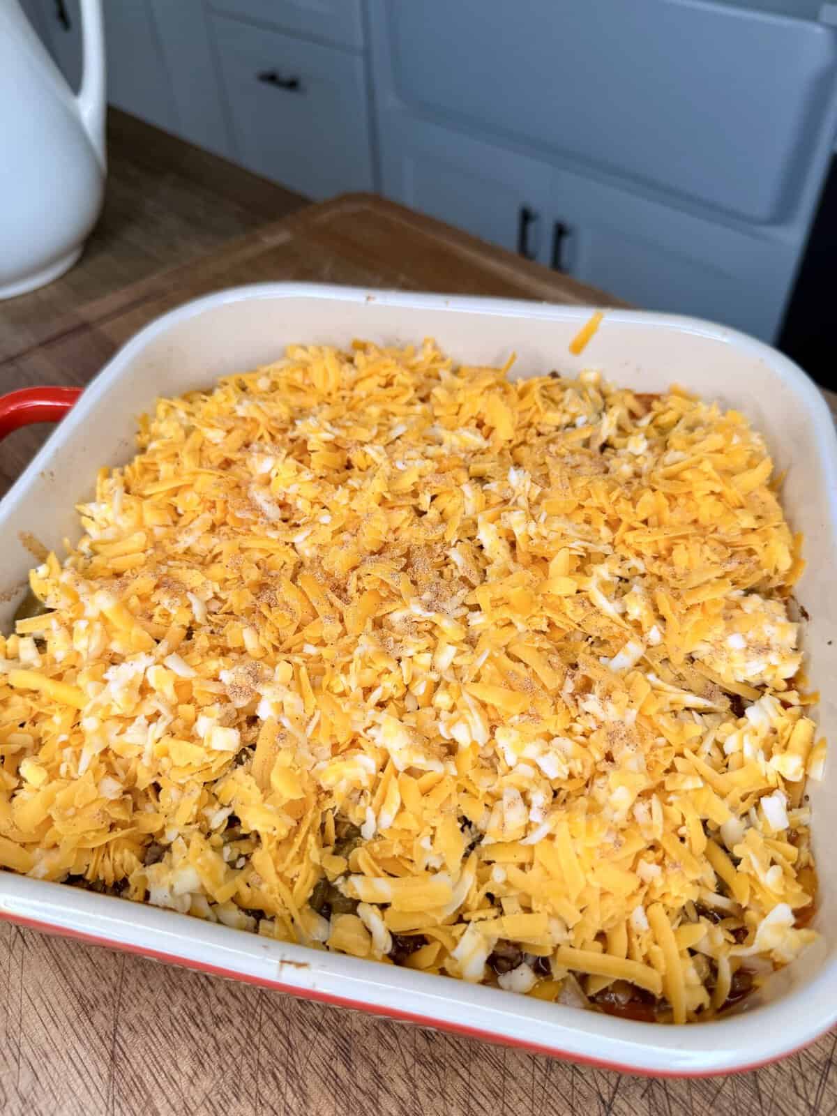 A square baking dish filled with a casserole topped with a layer of shredded cheddar cheese, sitting on a wooden countertop in a kitchen.