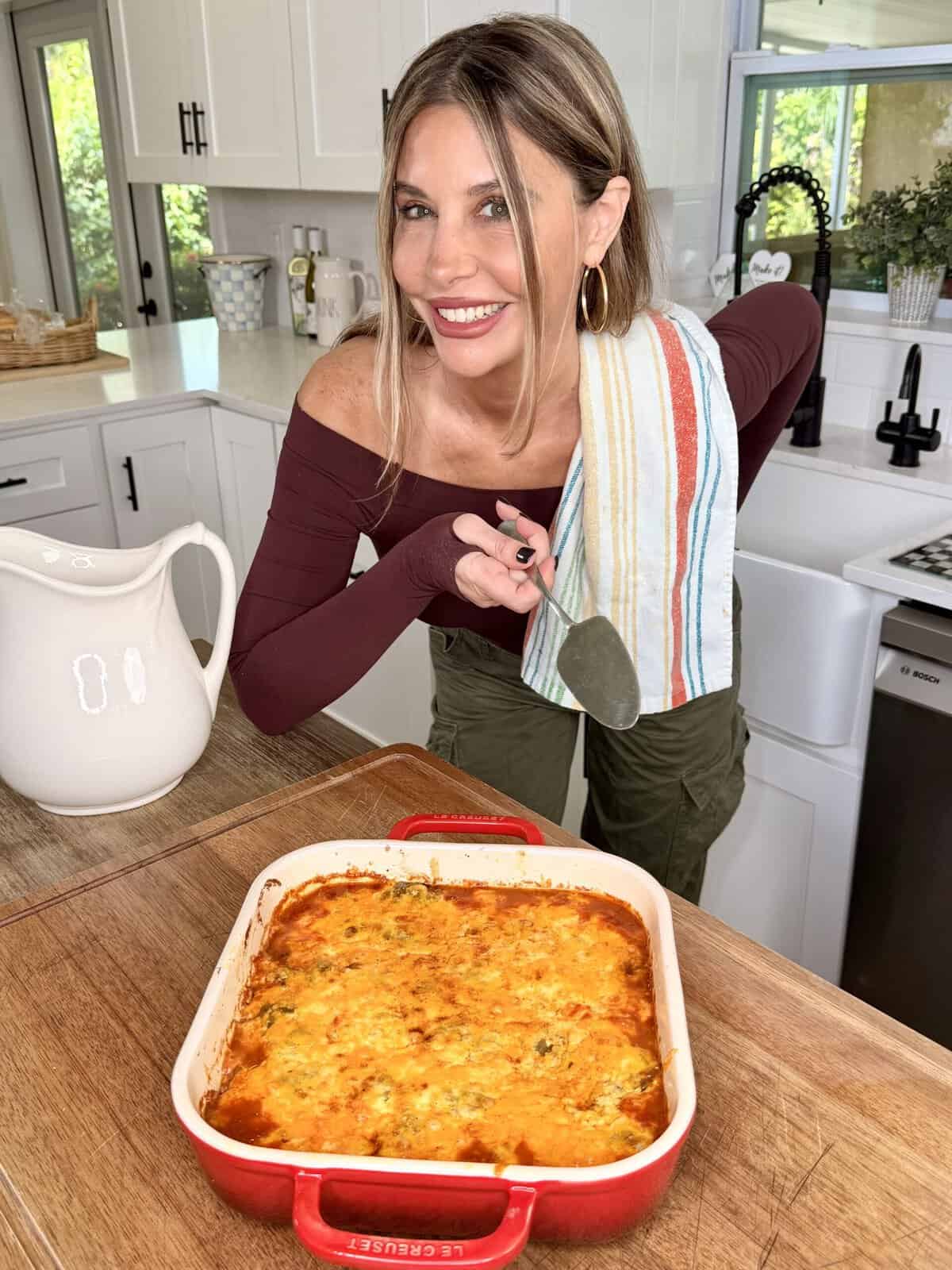 A woman in a kitchen smiles at the camera, holding a towel and a spatula beside a red baking dish filled with a baked casserole. White cabinets and a window are in the background.