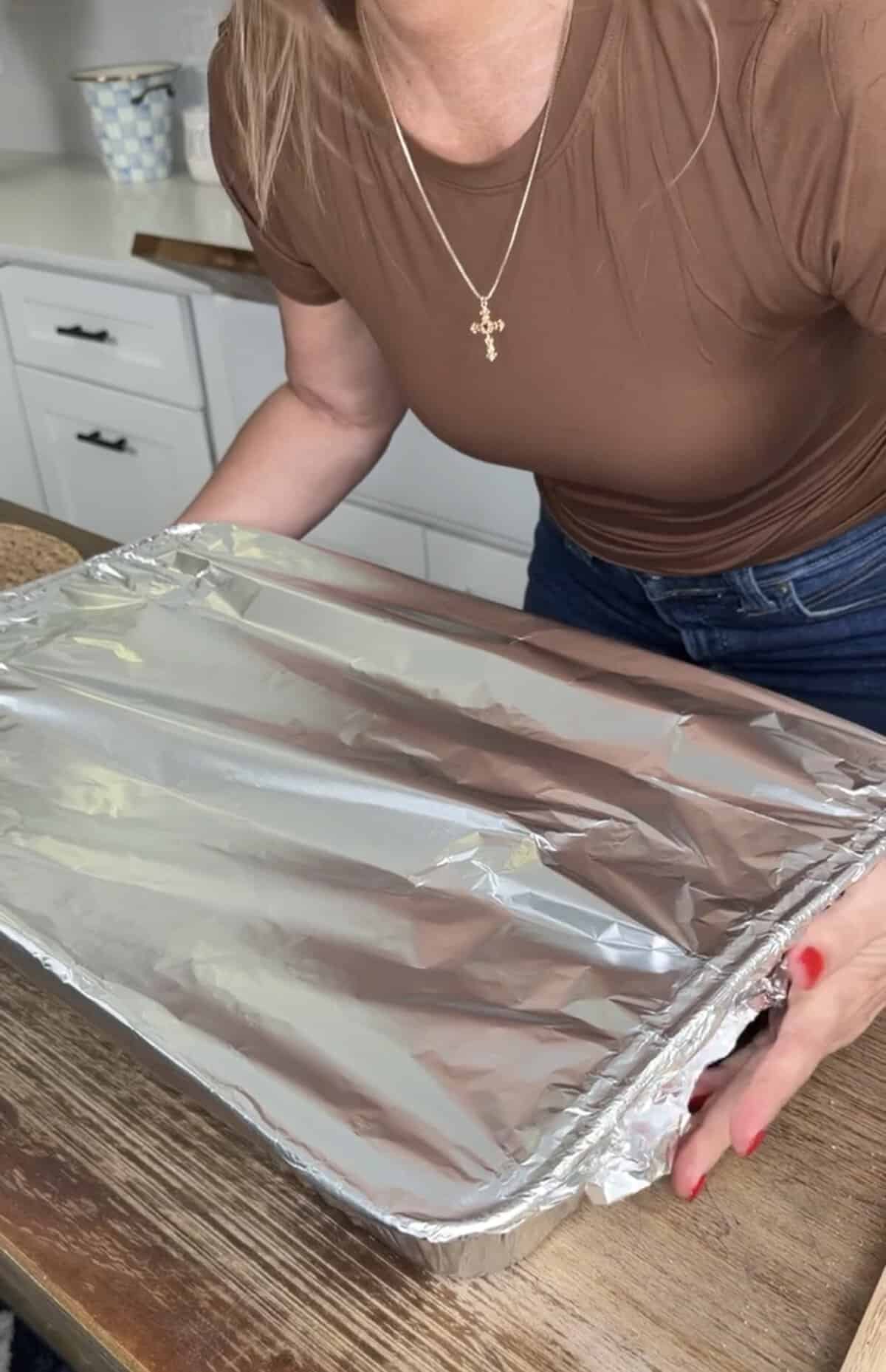 A person in a brown shirt and jeans holds a large rectangular tray covered with aluminum foil in a kitchen, preparing to place it on a wooden table.