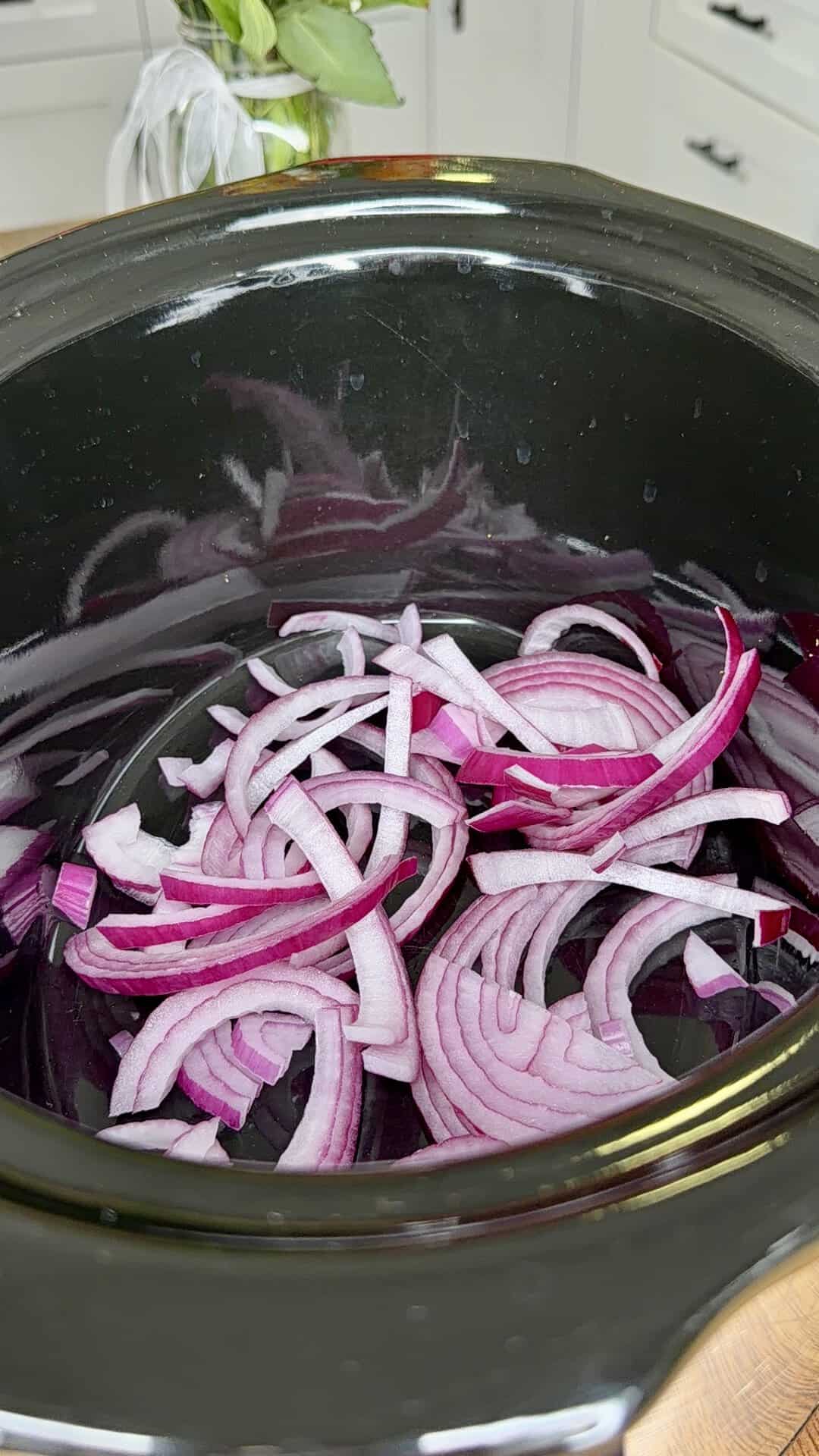 Thinly sliced red onions spread at the bottom of a black slow cooker, ready for cooking. A kitchen counter with a plant and drawers is visible in the background.