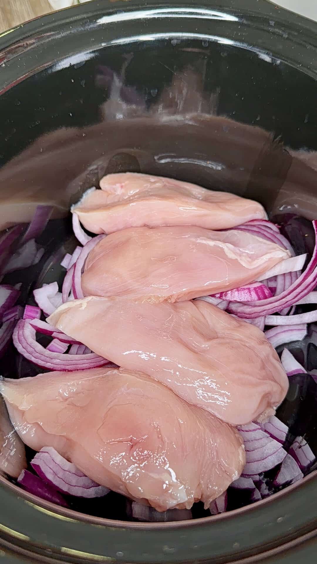 Raw chicken breasts placed on a bed of sliced red onions inside a black slow cooker, ready for cooking.