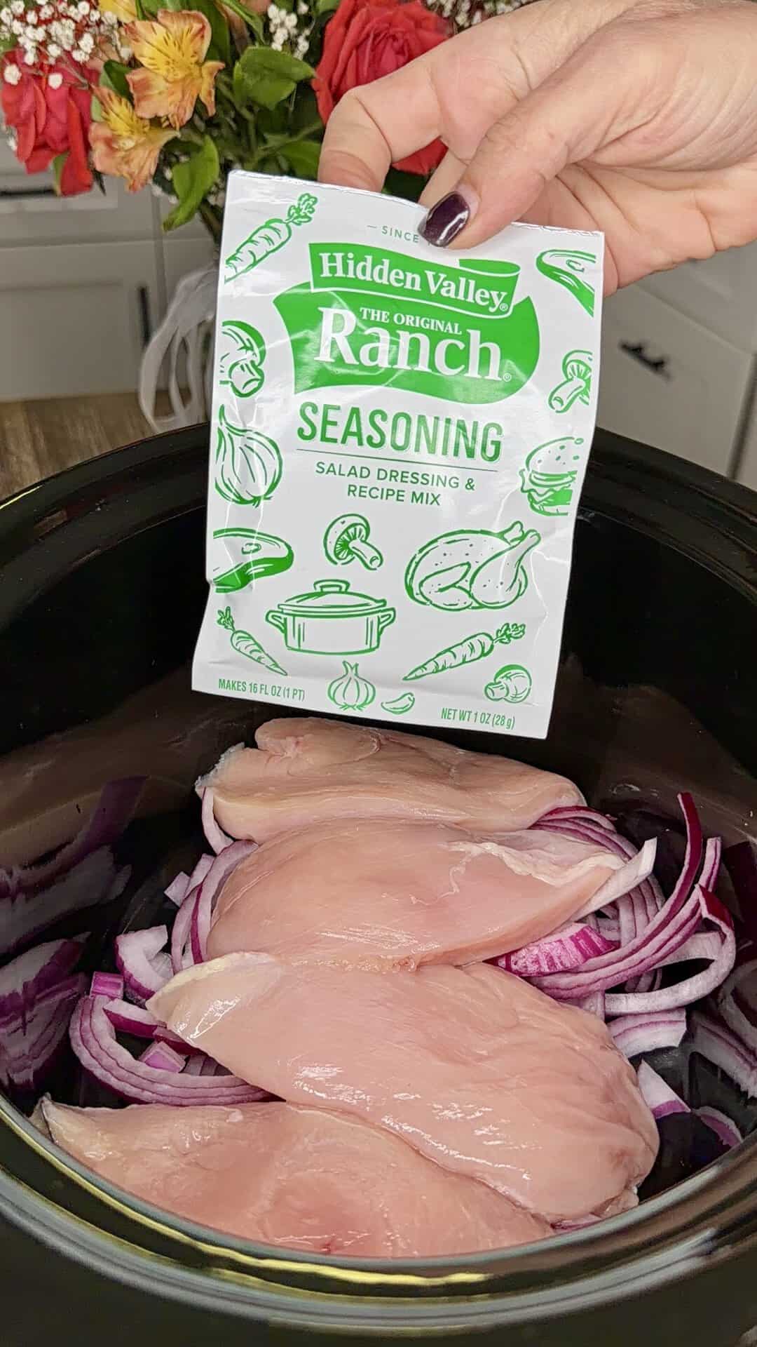 A hand holds a packet of Hidden Valley Ranch seasoning over a slow cooker containing raw chicken breasts and sliced red onions. A bouquet of flowers is visible in the background.