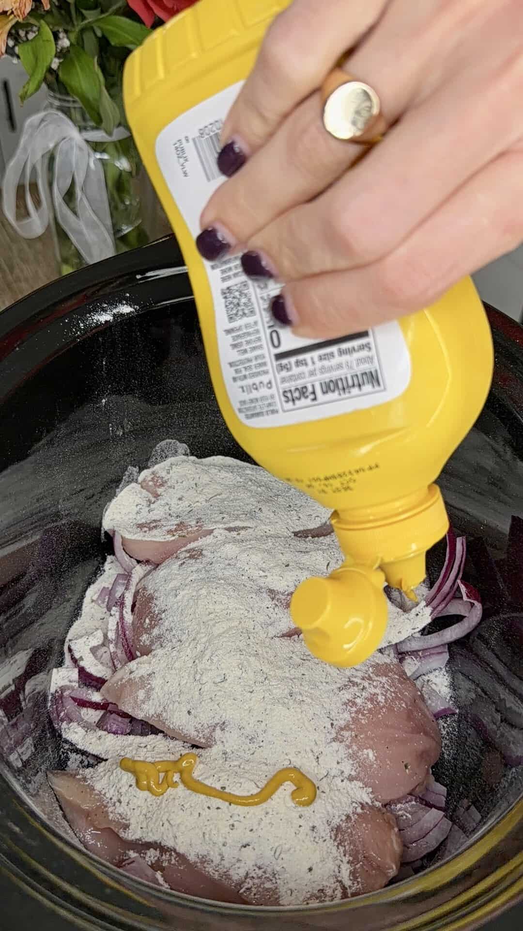 A hand squeezes yellow mustard onto raw chicken breasts topped with a powdered seasoning and sliced red onions inside a black slow cooker.