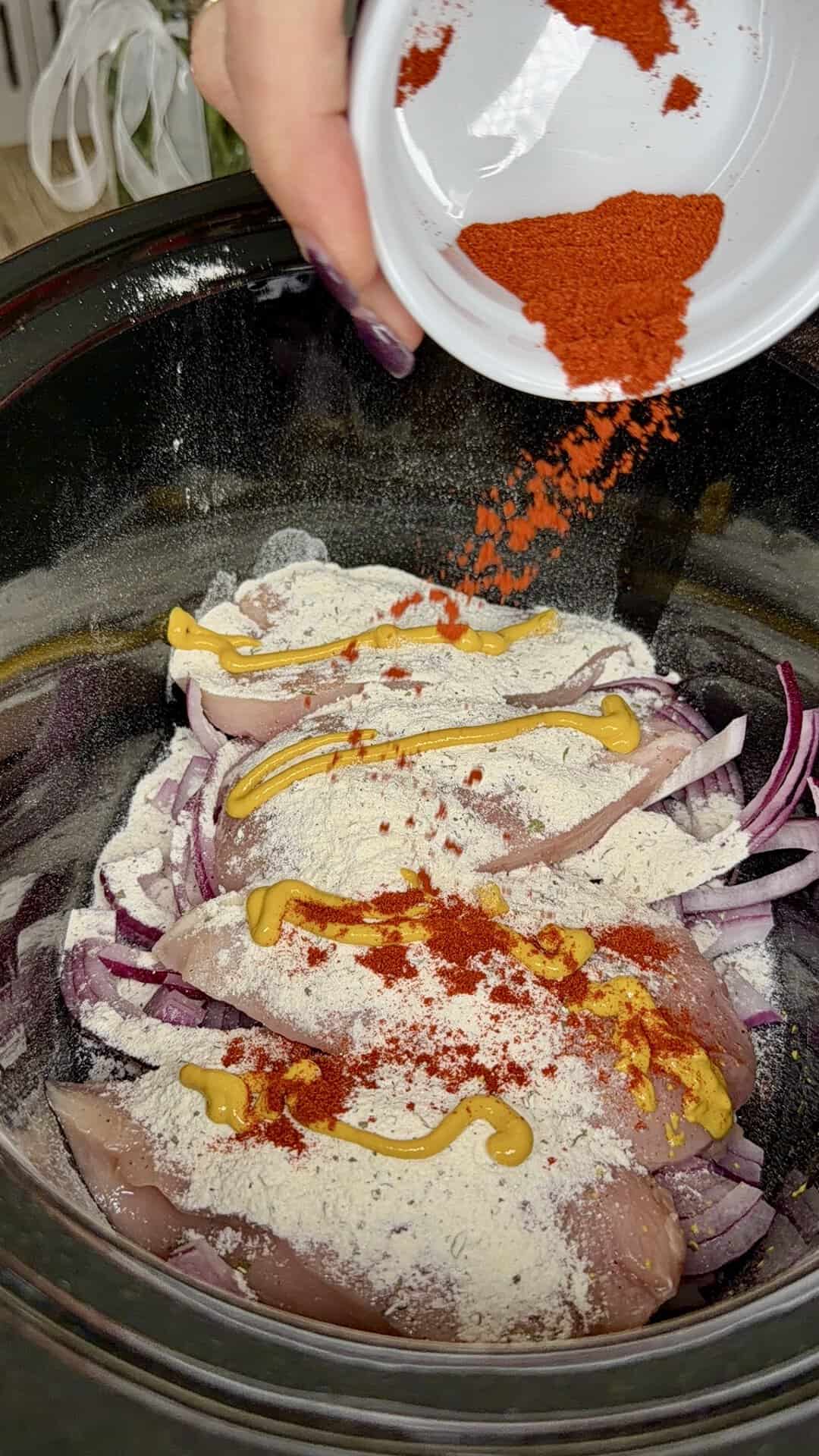 A hand sprinkles red paprika from a small white bowl over raw chicken breasts in a slow cooker. The chicken is topped with sliced red onions, mustard, and a dusting of white seasoning.