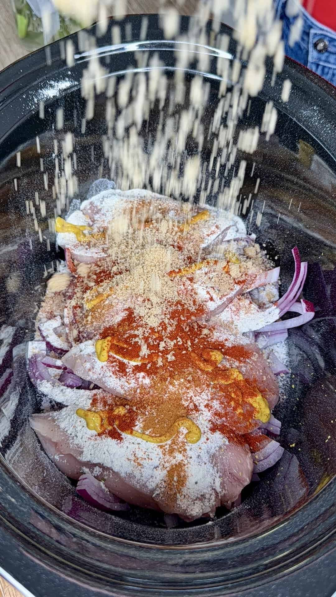 Raw chicken and sliced red onions in a slow cooker are topped with various spices and seasonings, while more seasoning is being sprinkled in from above.