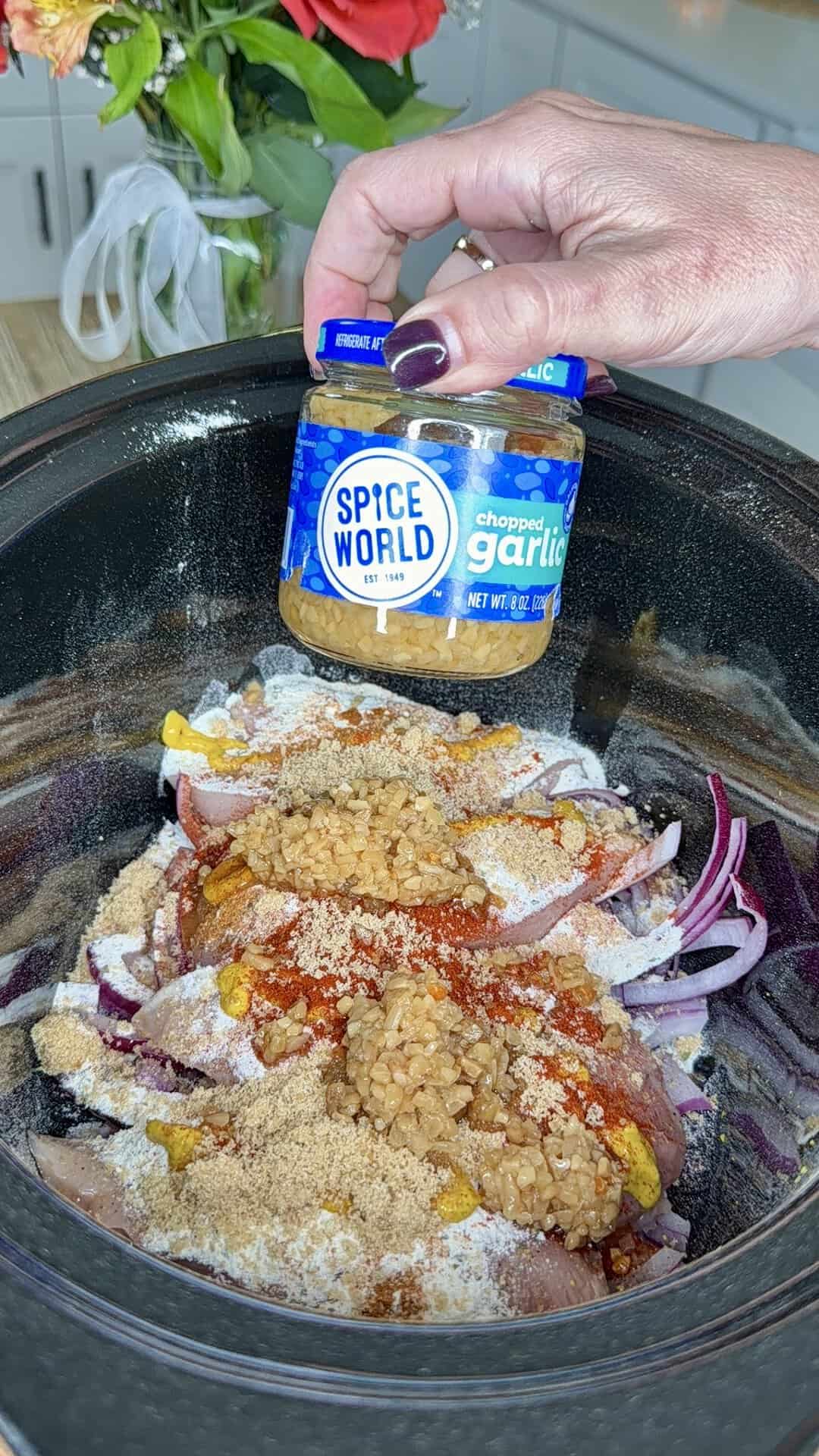 A hand holds a jar of Spice World chopped garlic above a slow cooker filled with raw chicken, red onions, and assorted spices. A bouquet of red flowers is visible in the background.