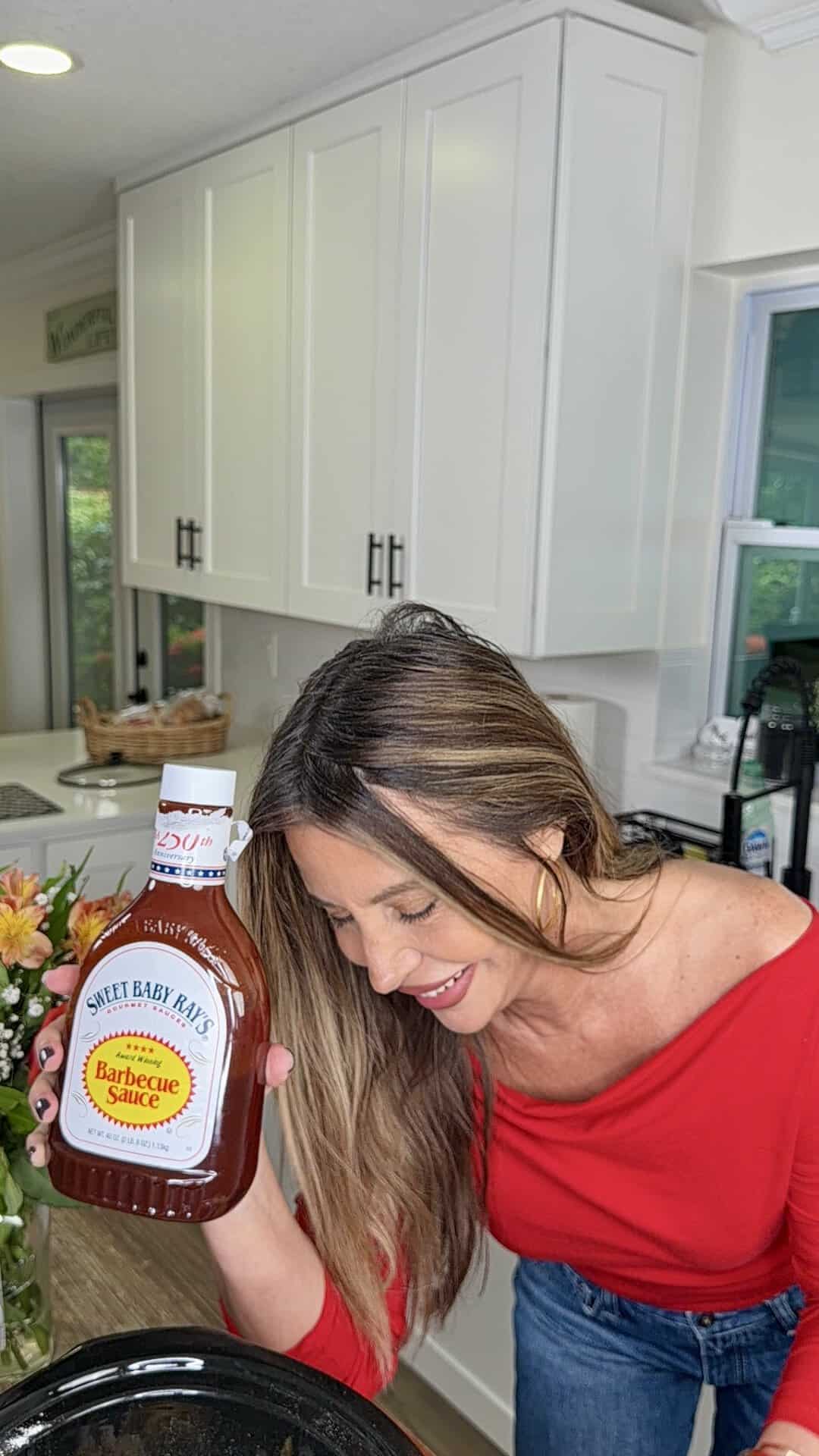 A woman in a red top smiles while holding a large bottle of Sweet Baby Rays Barbecue Sauce in a bright kitchen. She is leaning over a counter near a vase of flowers.