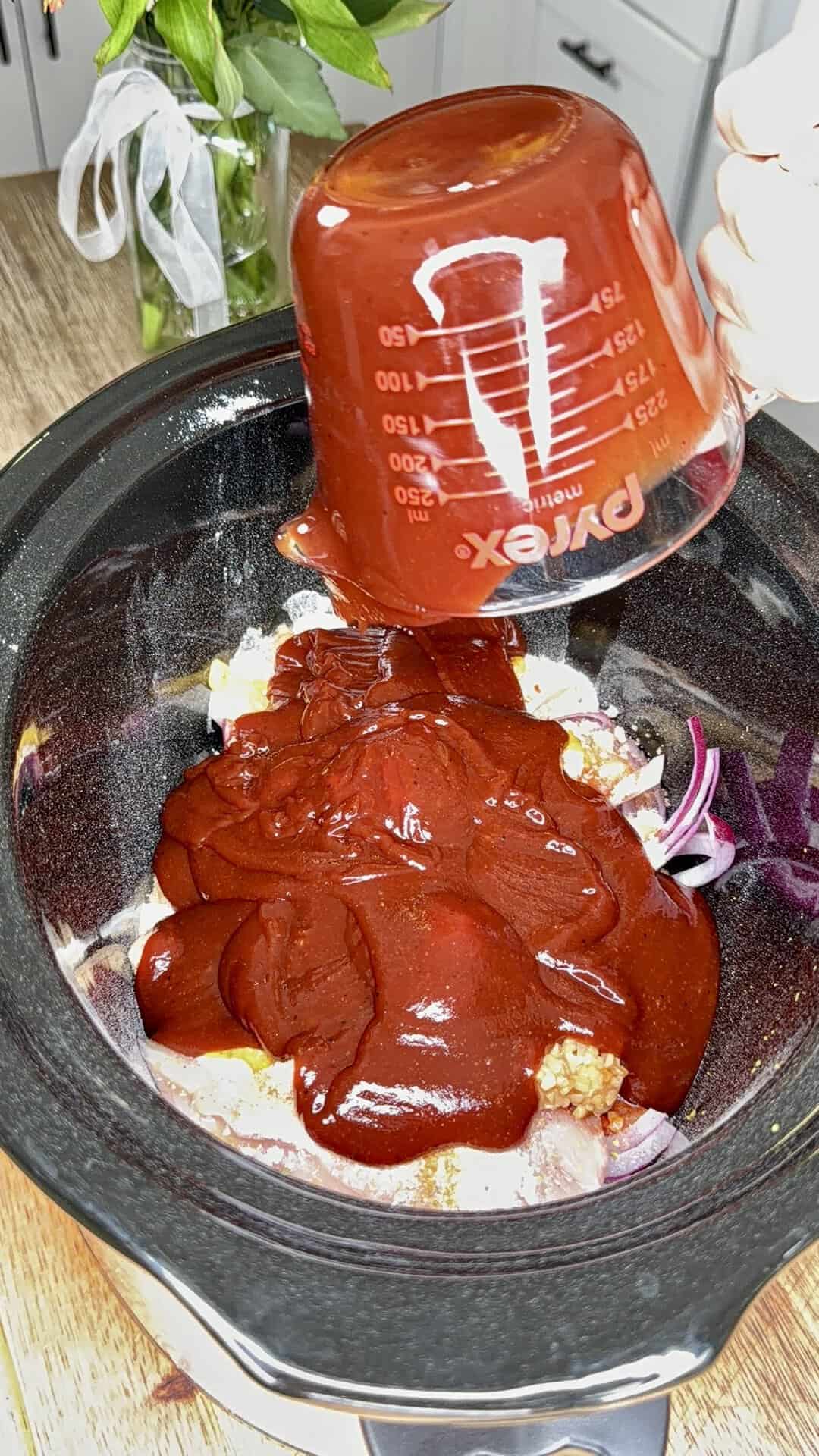 A hand pours a thick red sauce from a measuring cup into a slow cooker filled with raw chicken and sliced onions on a kitchen counter.
