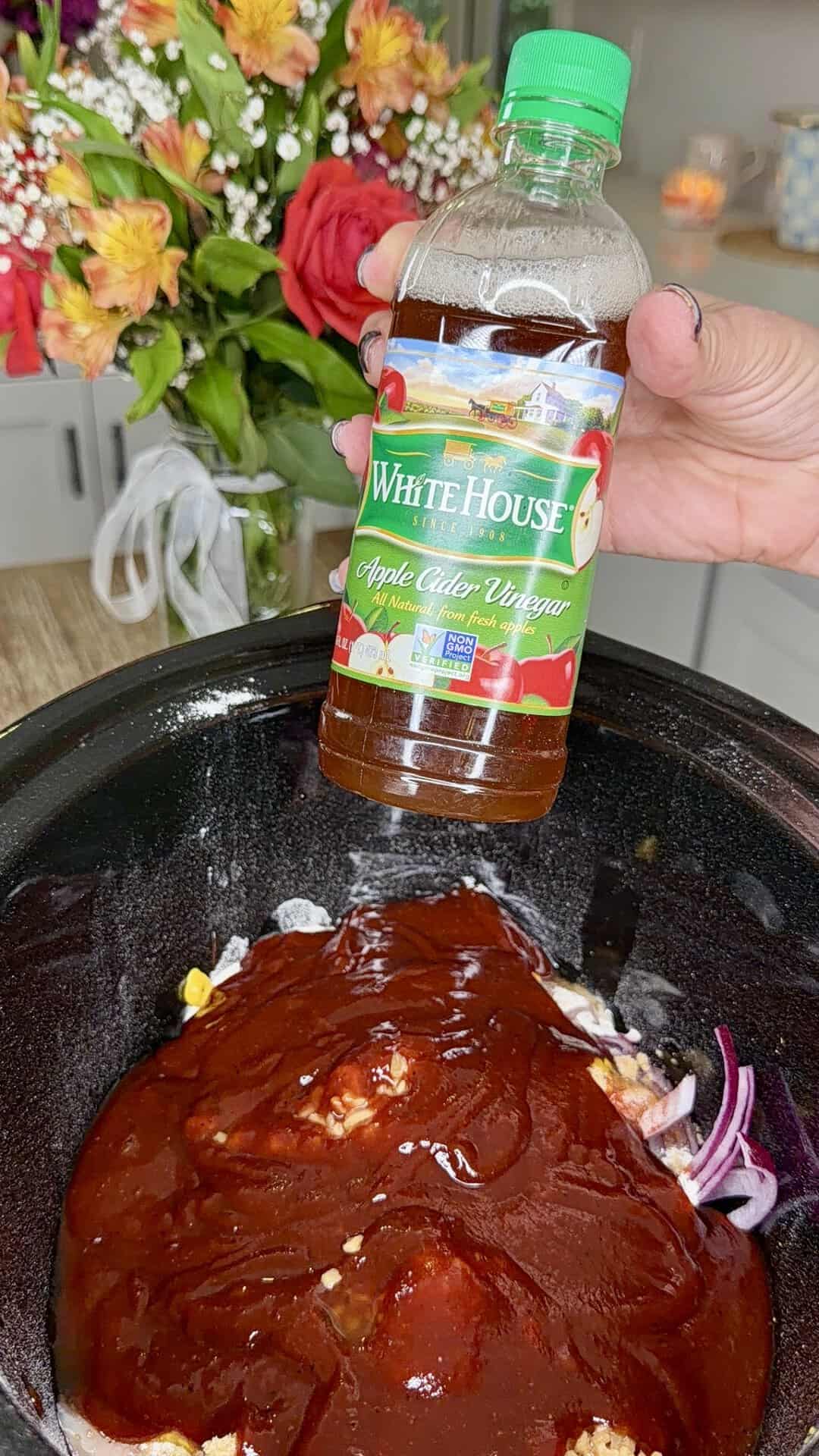 A hand holds a bottle of White House apple cider vinegar above a slow cooker filled with barbecue sauce, onions, and other ingredients. A vase of colorful flowers sits in the background on a kitchen counter.