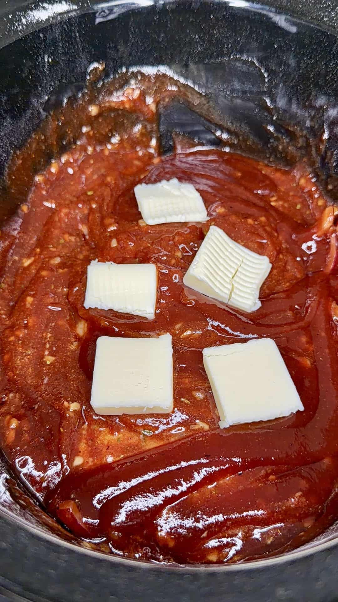 Close-up of a thick red sauce, possibly barbecue or tomato-based, in a black pot, topped with several small, rectangular slices of butter.