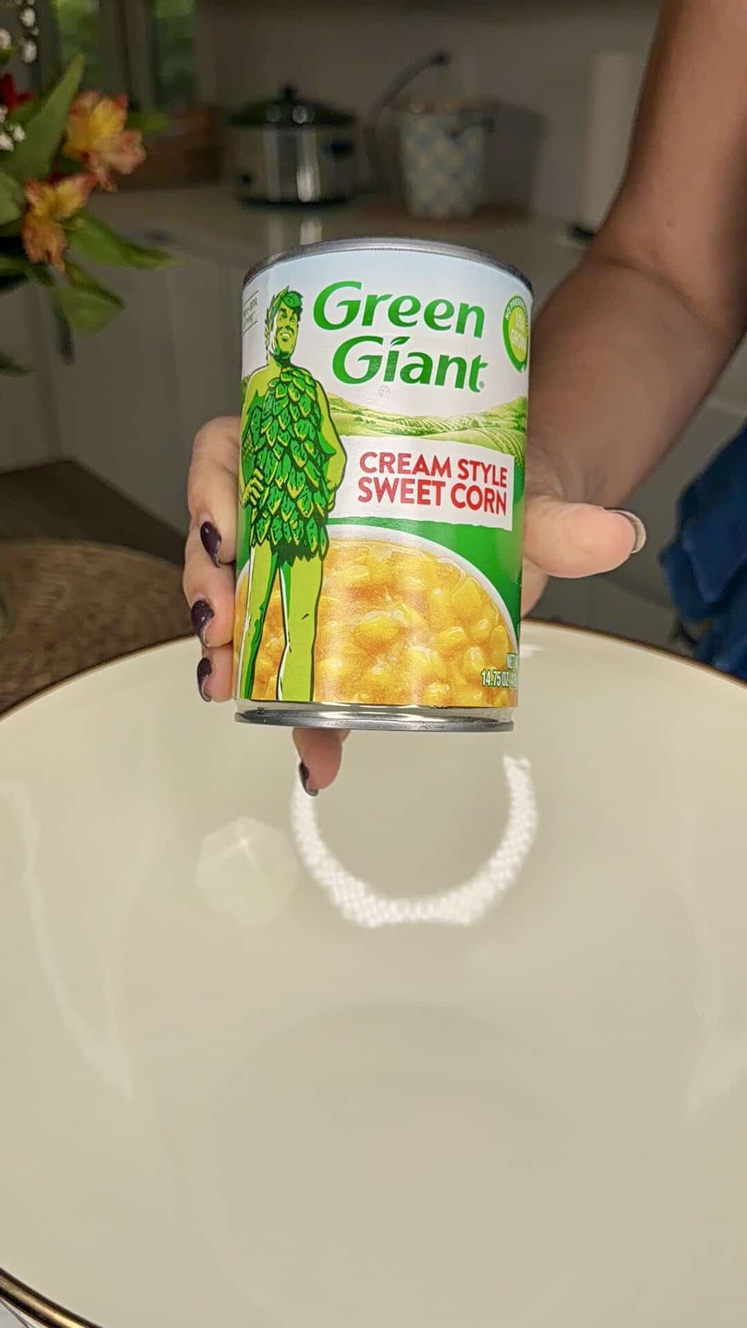 A hand holds a can of Green Giant Cream Style Sweet Corn over a large empty white bowl in a kitchen setting.