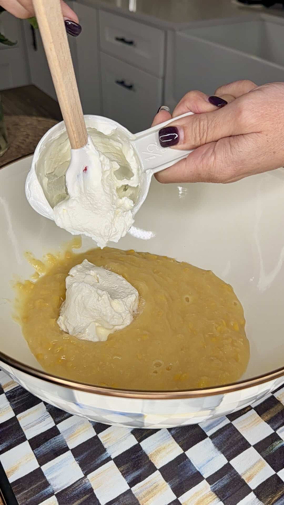 A person adds a scoop of sour cream from a measuring cup into a bowl of yellow cornbread batter using a wooden spatula. The bowl sits on a countertop with a checkered towel underneath.