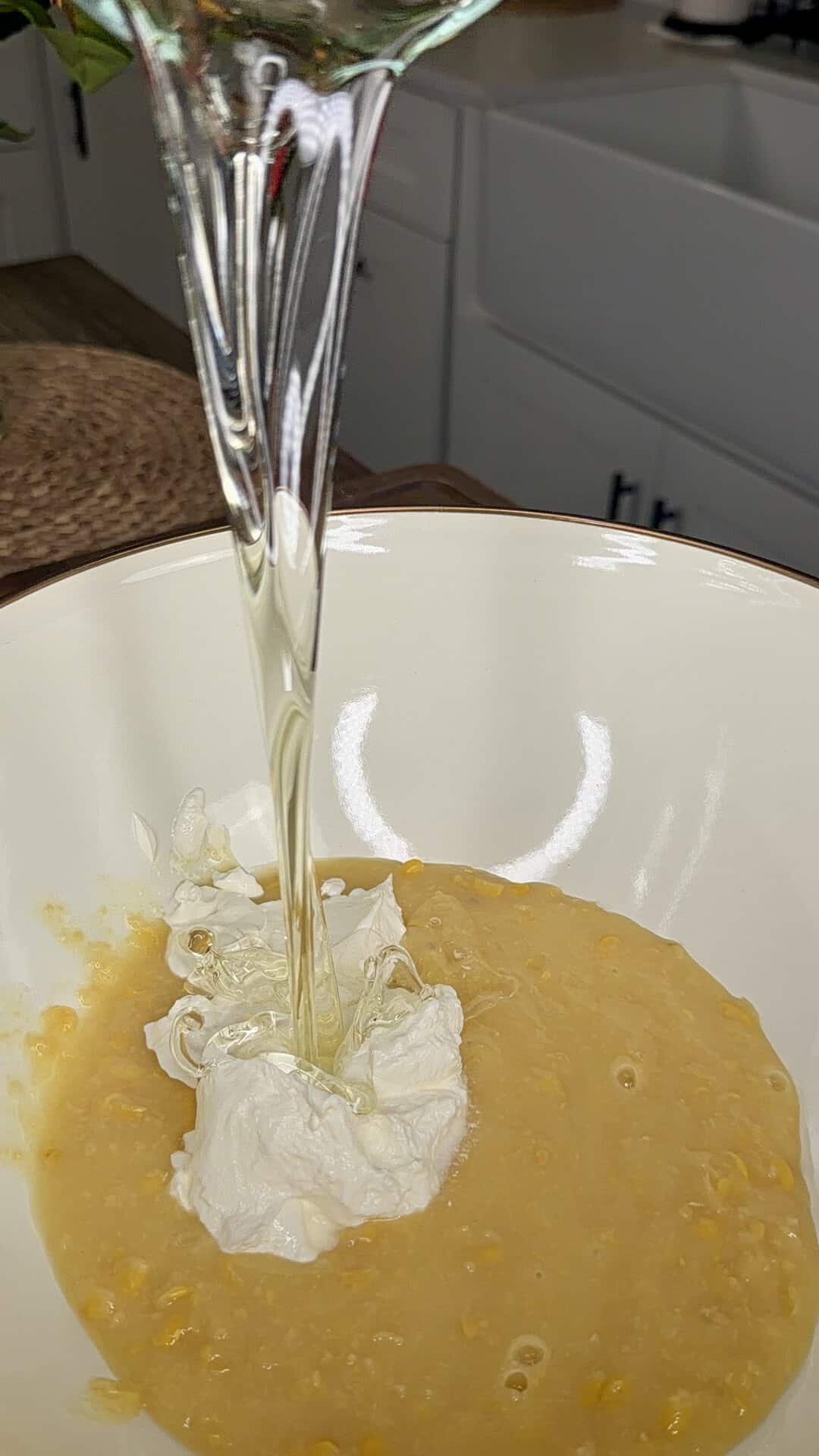 Clear oil is being poured into a bowl containing a mixture of creamed corn and a dollop of white cream cheese, set on a kitchen countertop with cabinets and a sink in the background.