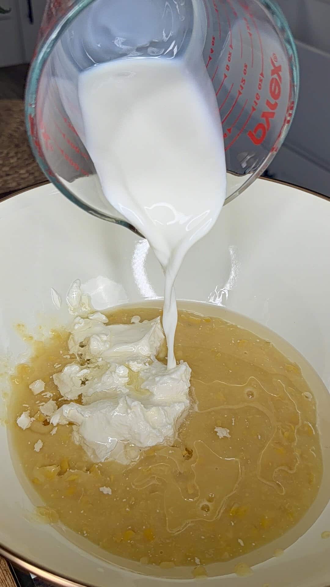 A glass measuring cup pours milk into a mixing bowl containing a yellowish batter and dollops of white cream cheese.