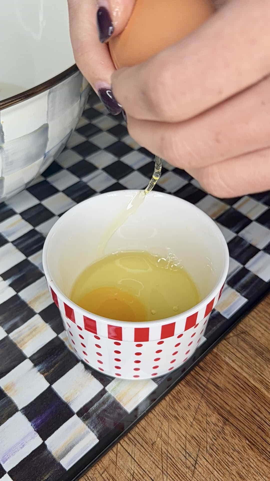 A person with dark-painted nails is cracking an egg into a white cup with red stripes, placed on a checkered surface next to a large metal bowl.