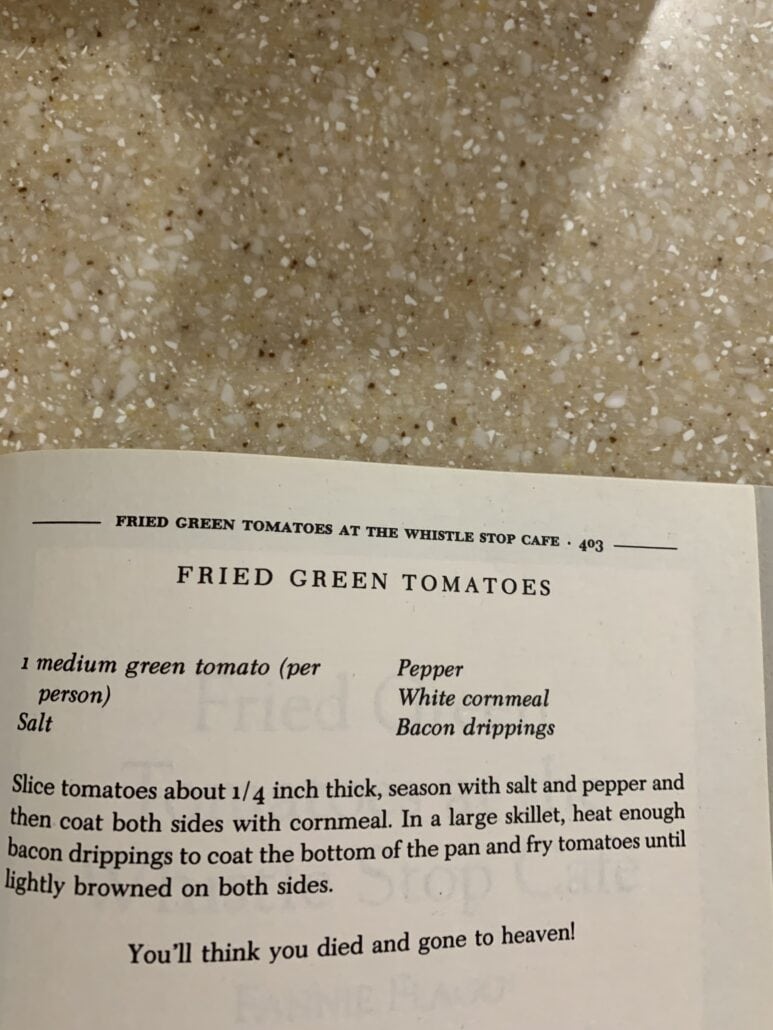 An open cookbook shows a fried green tomatoes recipe on a speckled countertop. The recipe lists ingredients and frying instructions, with a note at the bottom: “You’ll think you died and gone to heaven!”.
