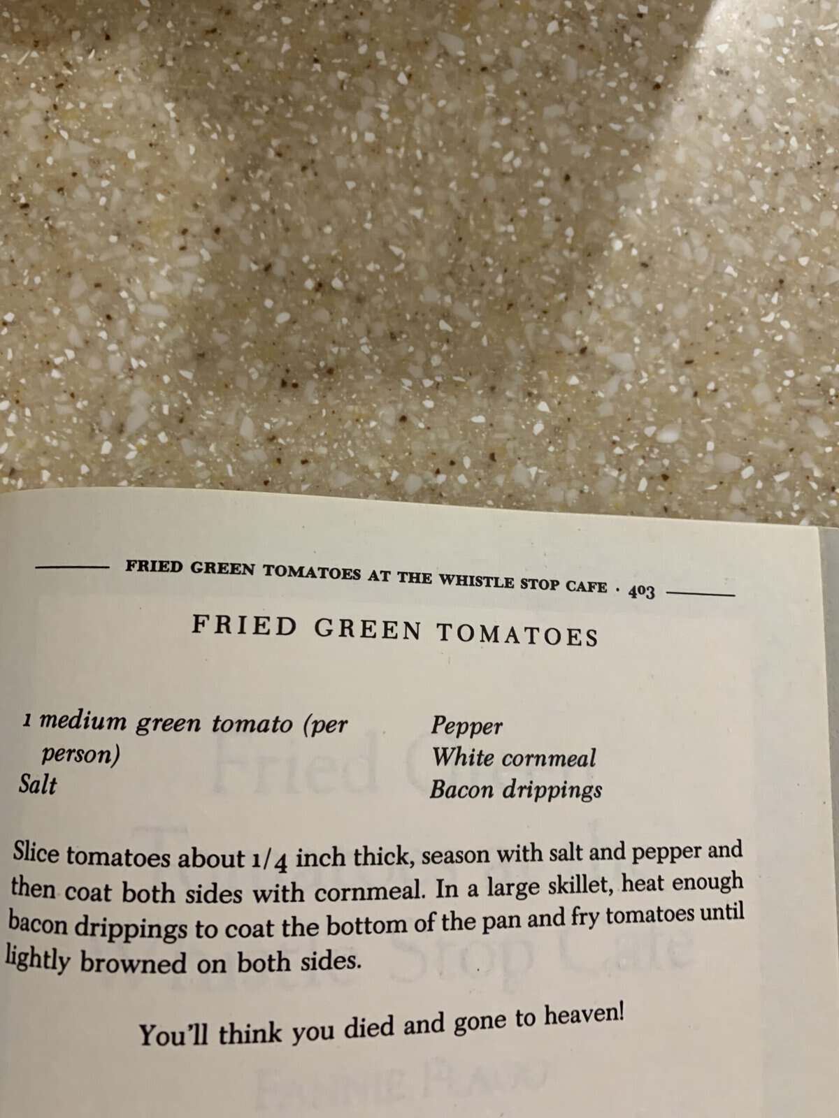 An open cookbook shows a fried green tomatoes recipe on a speckled countertop. The recipe lists ingredients and frying instructions, with a note at the bottom: “You’ll think you died and gone to heaven!”.