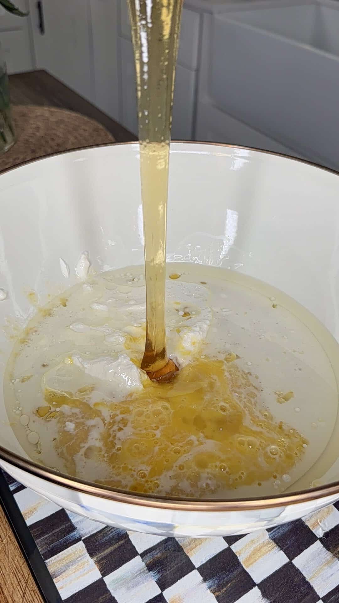 A stream of golden honey is being poured into a white bowl that already contains a creamy liquid, likely yogurt or milk, on a patterned surface in a kitchen setting.