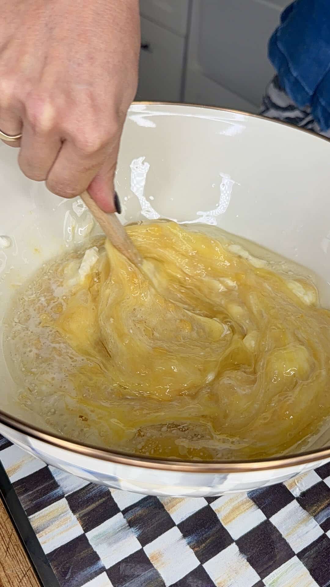 A hand stirs eggs and other ingredients in a large white bowl with a wooden spoon, mixing them together on a checkered surface.