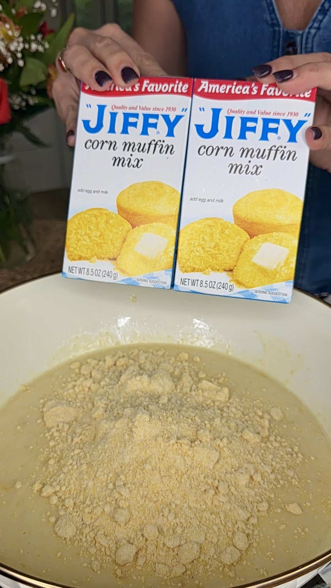 A person holds two boxes of Jiffy corn muffin mix above a bowl containing mixed batter and corn muffin mix powder, ready for further preparation.