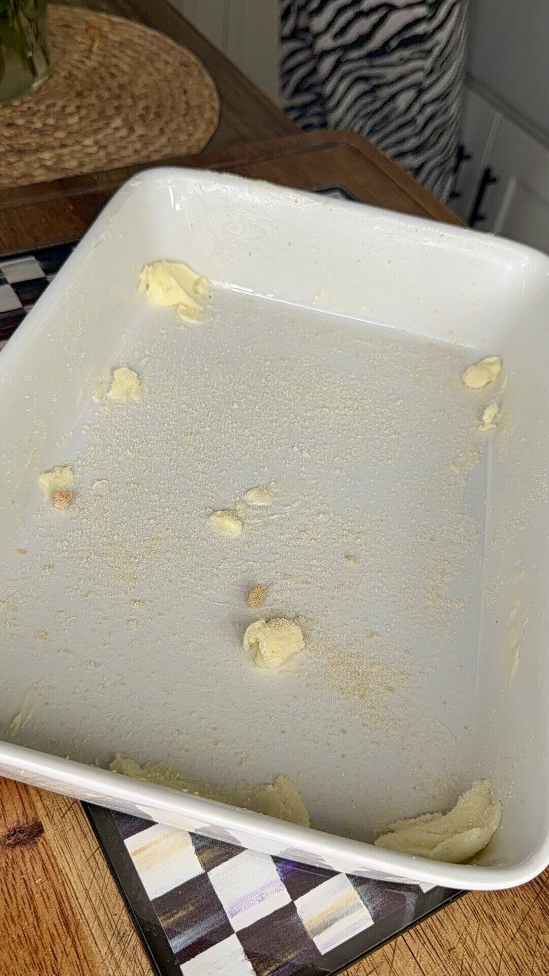 A white baking dish with small dollops of butter and a light dusting of breadcrumbs on the bottom, sitting on a wooden countertop with a woven placemat and patterned trivet nearby.