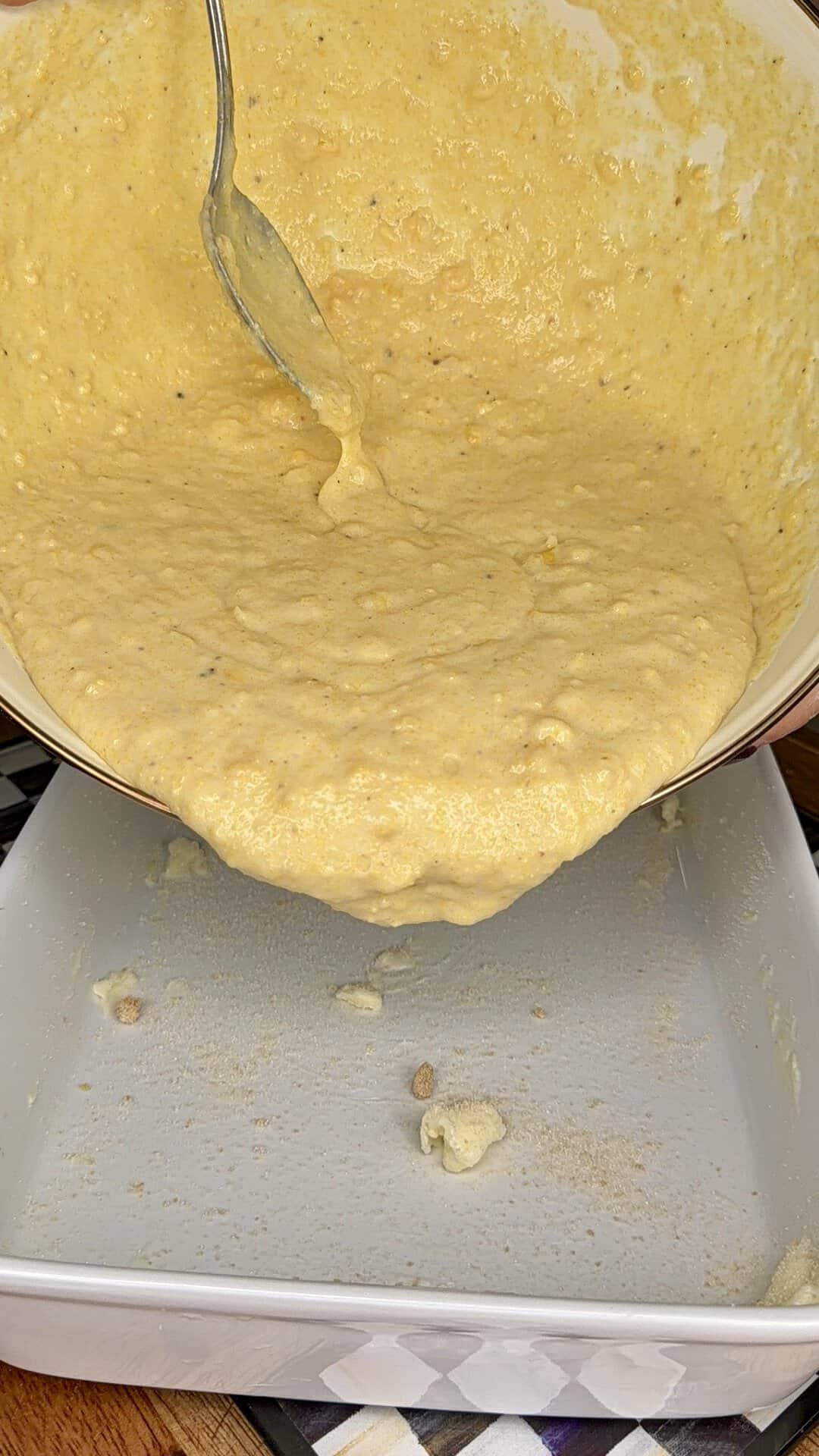 A hand holds a bowl, pouring thick yellow cornbread batter with a fork into a rectangular white baking dish. Some batter has already dripped into the dish.