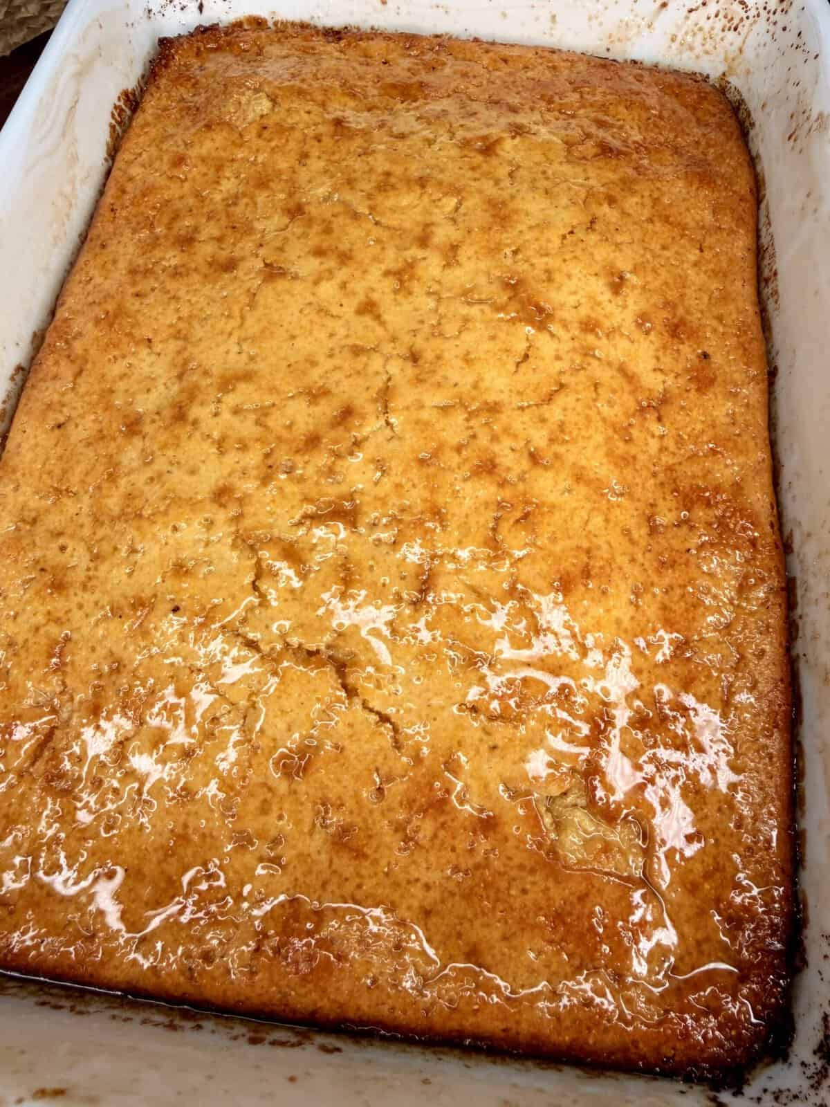 A freshly baked, golden-brown cake in a white rectangular baking dish, with a shiny glaze on top and slightly crispy edges.