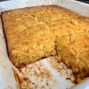 A golden-brown cornbread with a moist, crumbly texture is shown in a white baking dish, with a square piece removed from one corner. The kitchen counter is visible in the background.