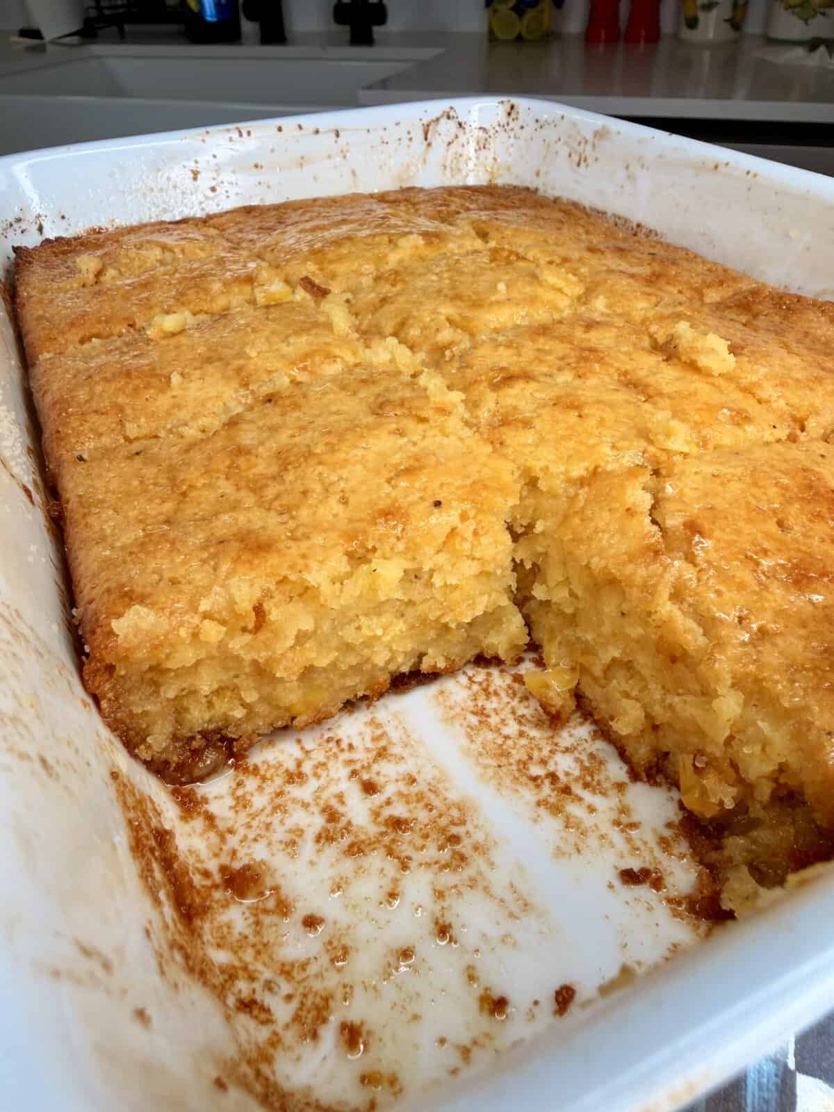 A golden-brown cornbread with a moist, crumbly texture is shown in a white baking dish, with a square piece removed from one corner. The kitchen counter is visible in the background.