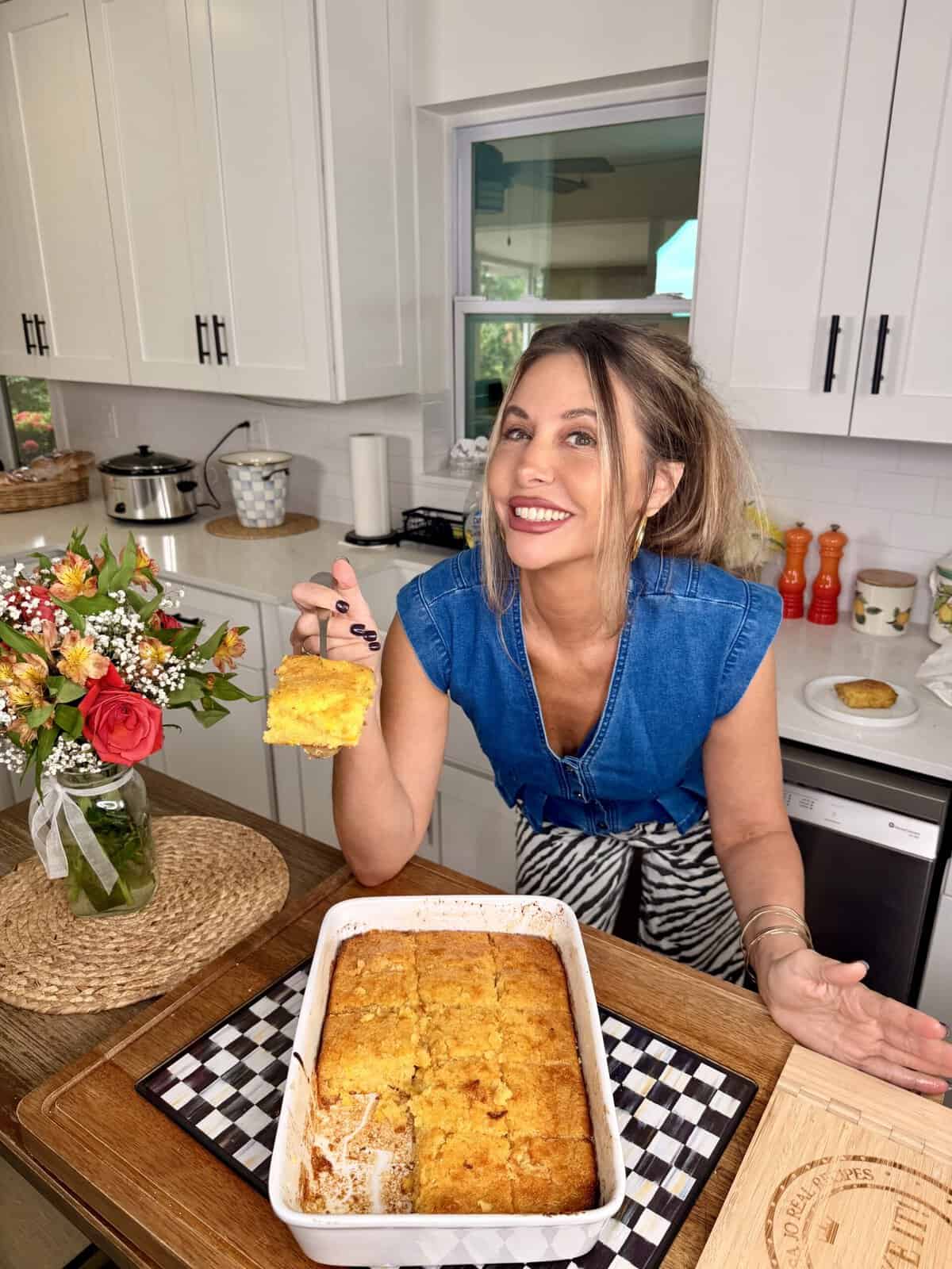 A smiling woman in a blue top holds up a serving of baked casserole in a kitchen. The casserole dish is on a wooden tray, and there are flowers and kitchen items in the background.
