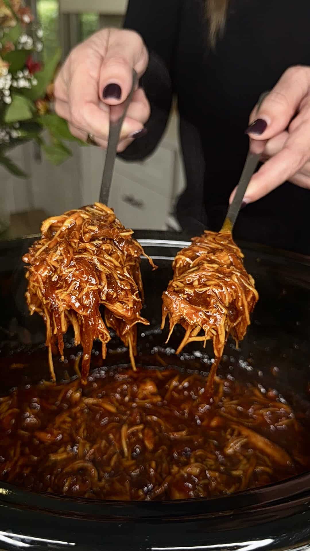 A person with dark nail polish uses two forks to lift saucy shredded barbecue chicken from a black slow cooker in a kitchen setting.