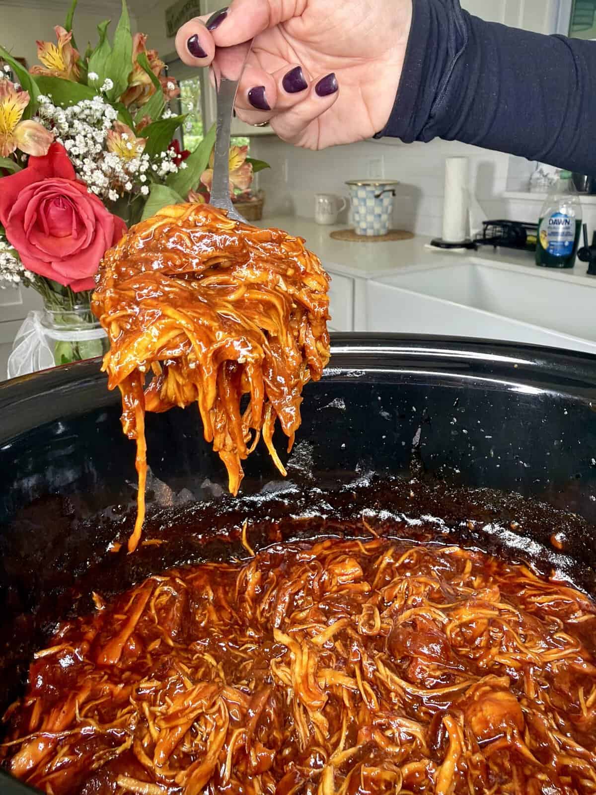 A hand with dark purple nails lifts a serving of saucy shredded barbecue chicken from a slow cooker; a bouquet of flowers and a kitchen counter are visible in the background.