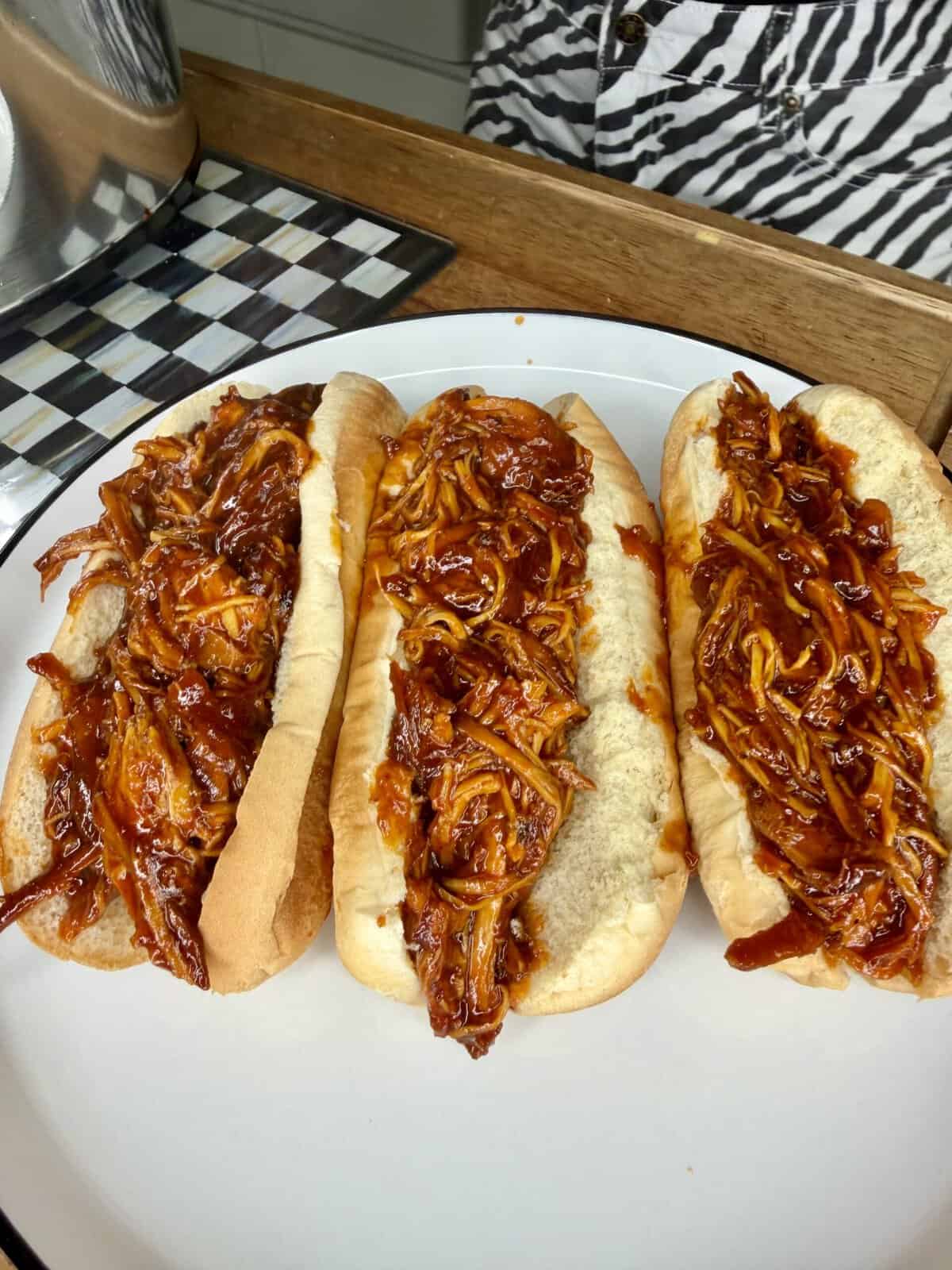 Three hot dog buns filled with shredded barbecue chicken sit on a white plate. The buns are lined up side by side, and the shredded chicken is coated in a rich, red barbecue sauce.
