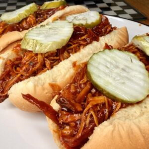 Three hot dog buns filled with shredded barbecue chicken and topped with sliced pickles are arranged on a white plate. A checkered tray and slow cooker are visible in the background.