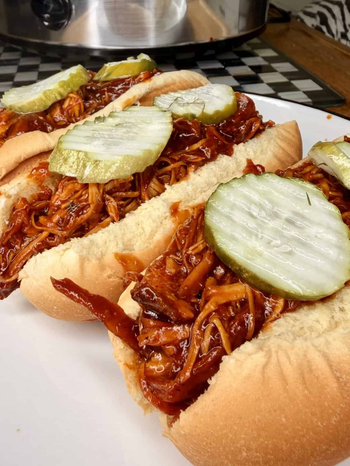 Three hot dog buns filled with shredded barbecue chicken and topped with sliced pickles are arranged on a white plate. A checkered tray and slow cooker are visible in the background.