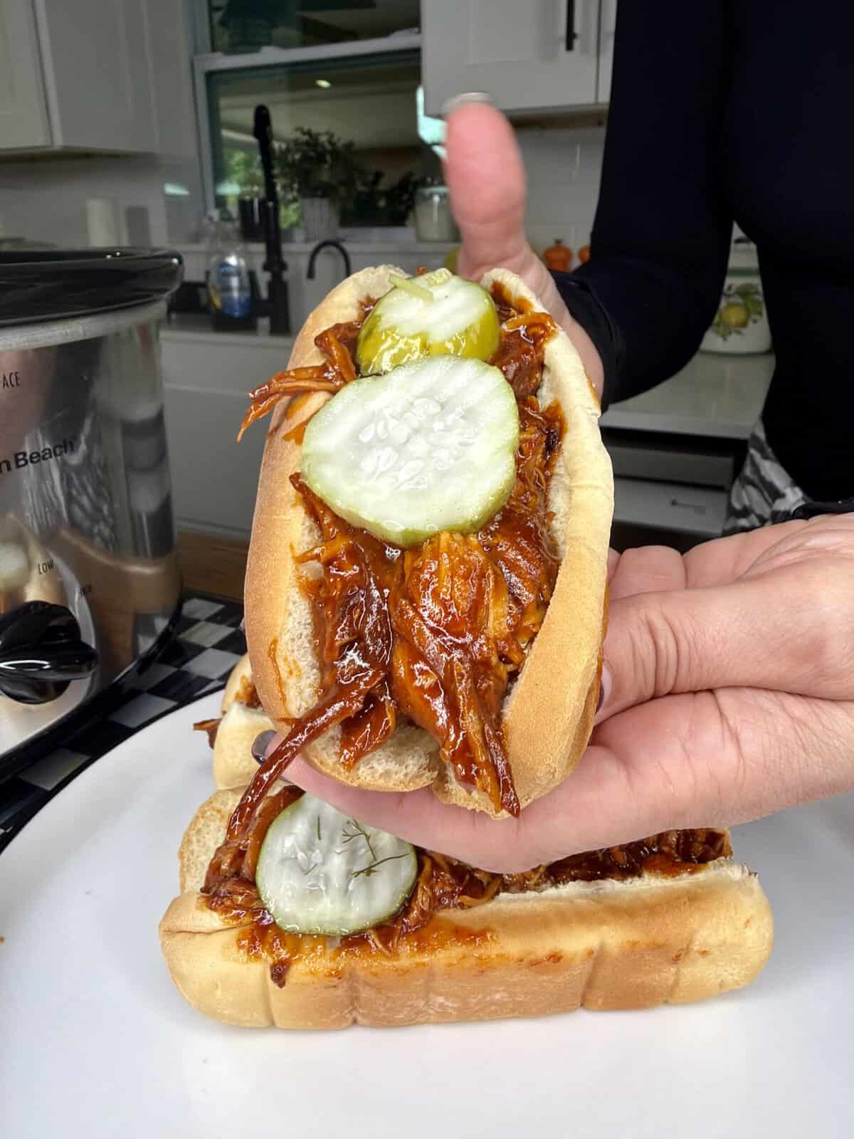A hand holds a barbecue pulled pork sandwich topped with pickle slices in a hot dog bun, with another similar sandwich on a plate in a kitchen setting.