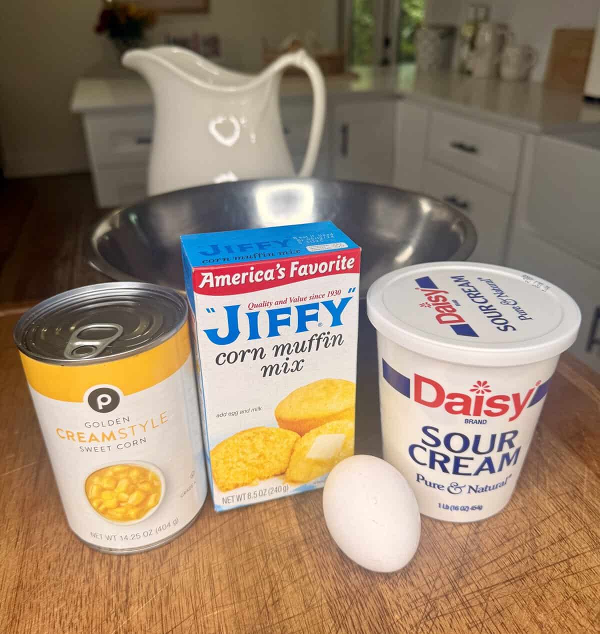 A can of cream style corn, Jiffy corn muffin mix, a tub of sour cream, and an egg sit on a kitchen counter with a large metal bowl and a white pitcher in the background.