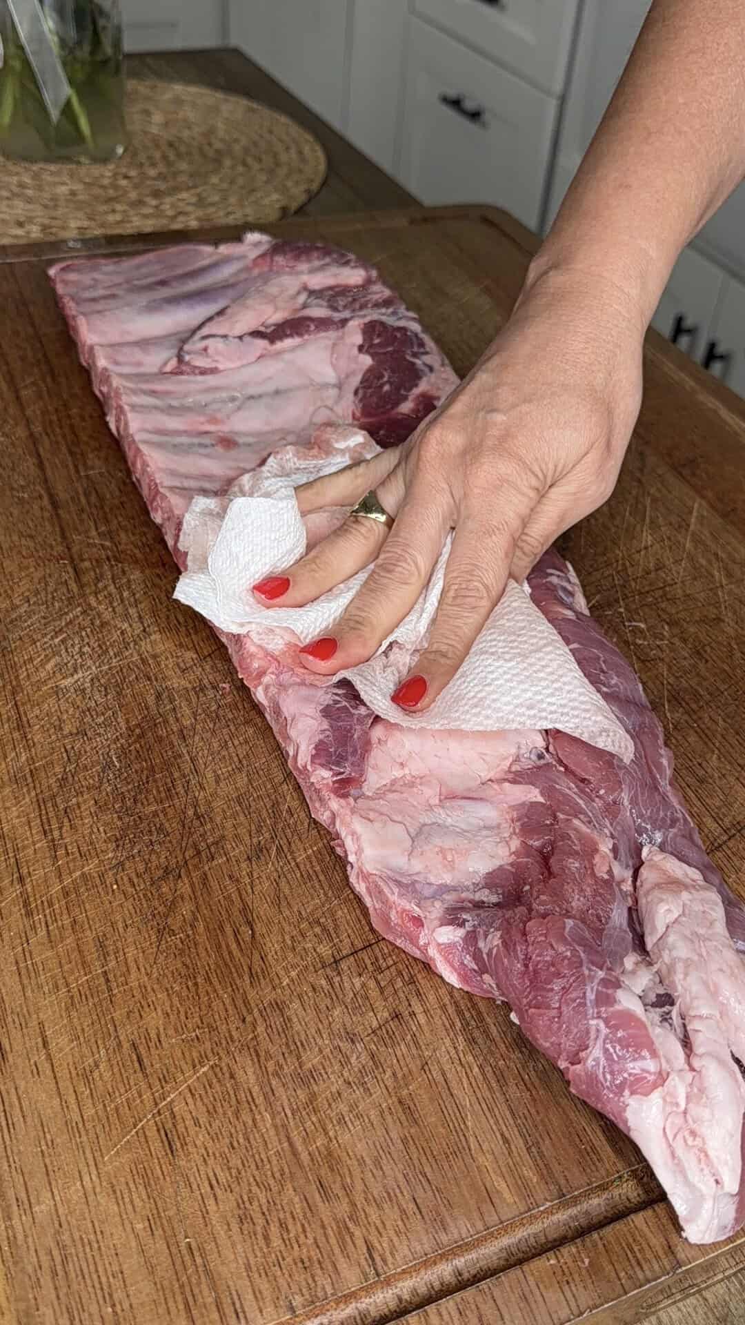 A hand with red nail polish uses a paper towel to pat dry a large rack of raw ribs on a wooden cutting board in a kitchen.
