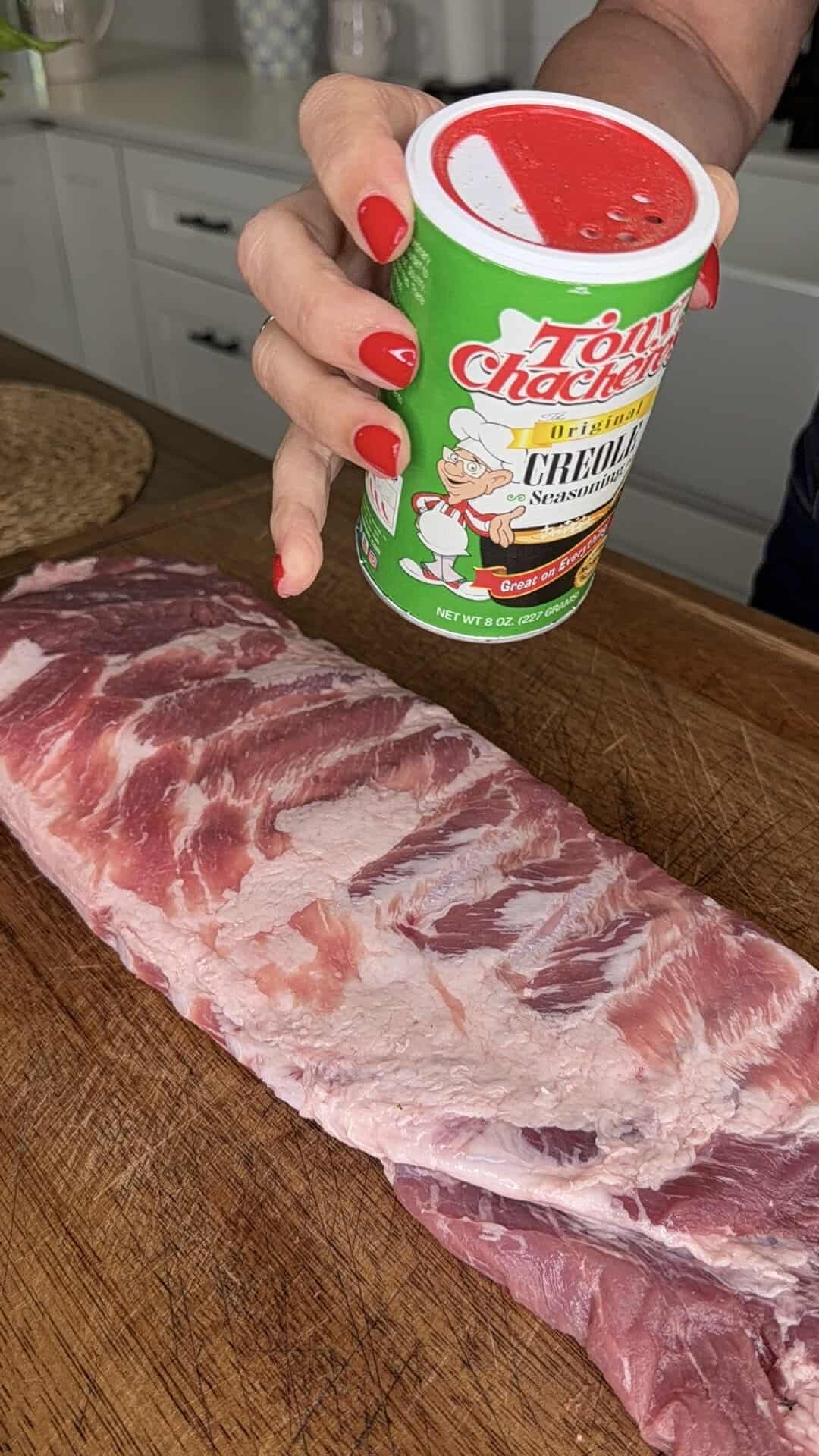 A hand with red nail polish holds a can of Tony Chachere’s Creole seasoning over a raw slab of pork ribs on a wooden cutting board in a kitchen.