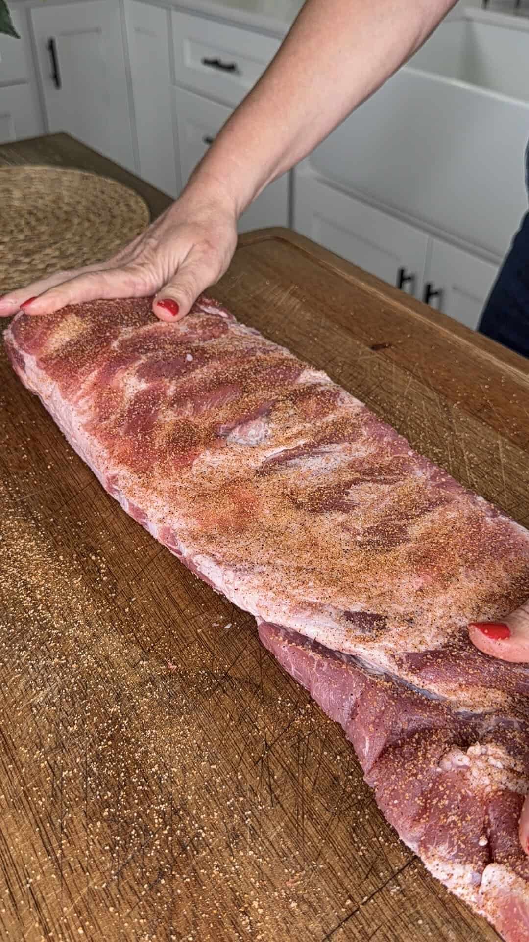 A person with red nail polish seasons a rack of raw pork ribs with dry rub on a wooden cutting board in a kitchen.