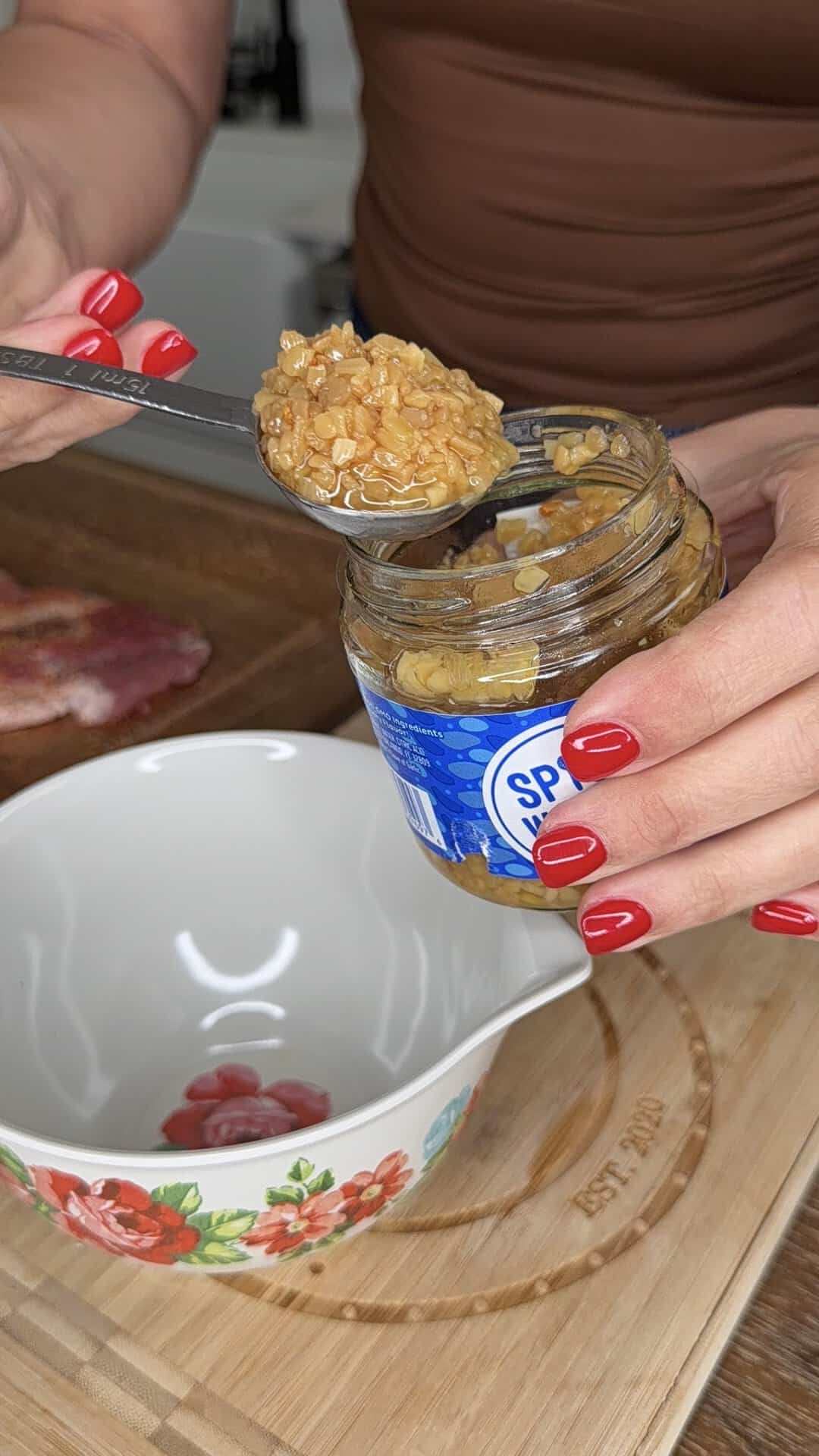 A person with red nail polish scoops minced garlic from a glass jar with a spoon over a white bowl decorated with red flowers, on a wooden cutting board.