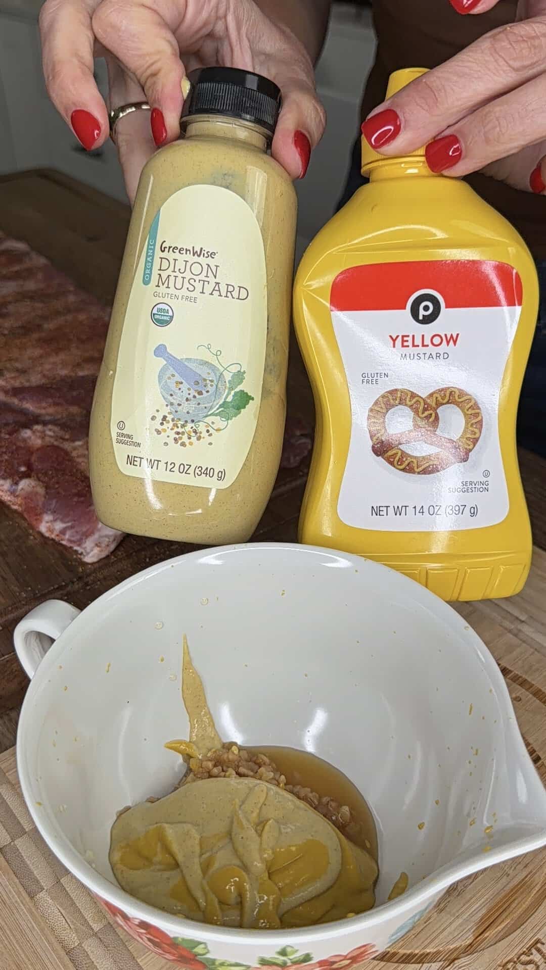 A person with red nail polish holds bottles of dijon and yellow mustard above a bowl containing both mustards. Raw meat is visible on a cutting board in the background.