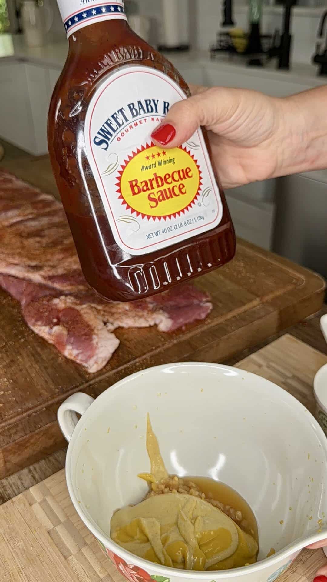 A hand holds a bottle of Sweet Baby Rays Barbecue Sauce over a kitchen counter with raw ribs on a cutting board and a bowl containing mustard and sauce in the foreground.