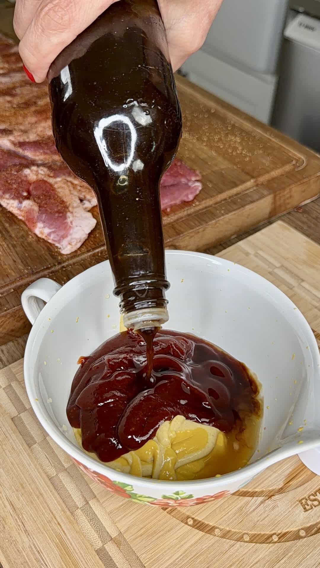 A hand pours barbecue sauce from a bottle into a white bowl containing mustard and honey, with seasoned raw meat and a cutting board in the background.