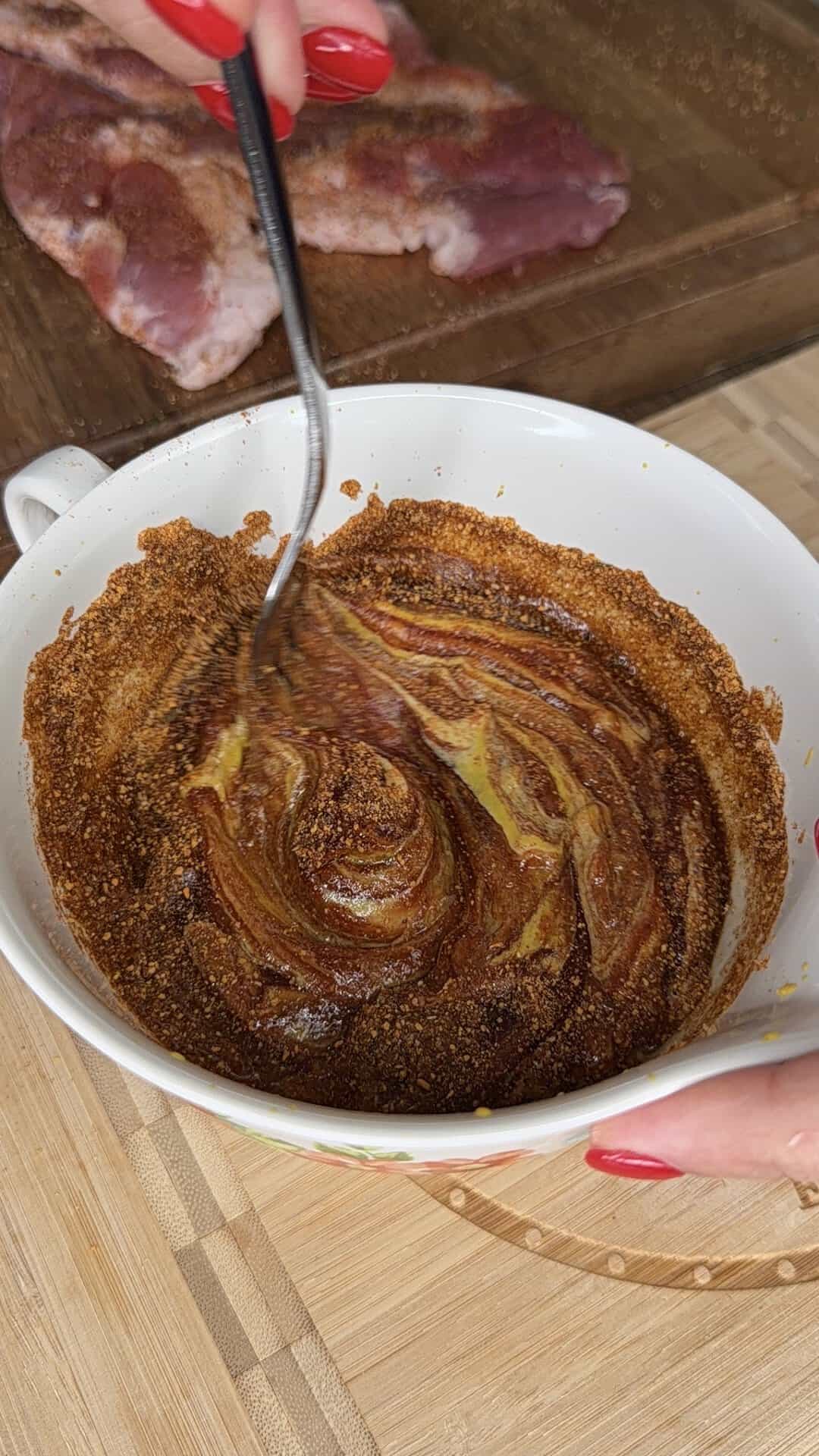 A person with red nails mixes a brown spice blend and yellow mustard in a white bowl with a fork. Raw meat sits on a cutting board in the background, placed on a wooden surface.