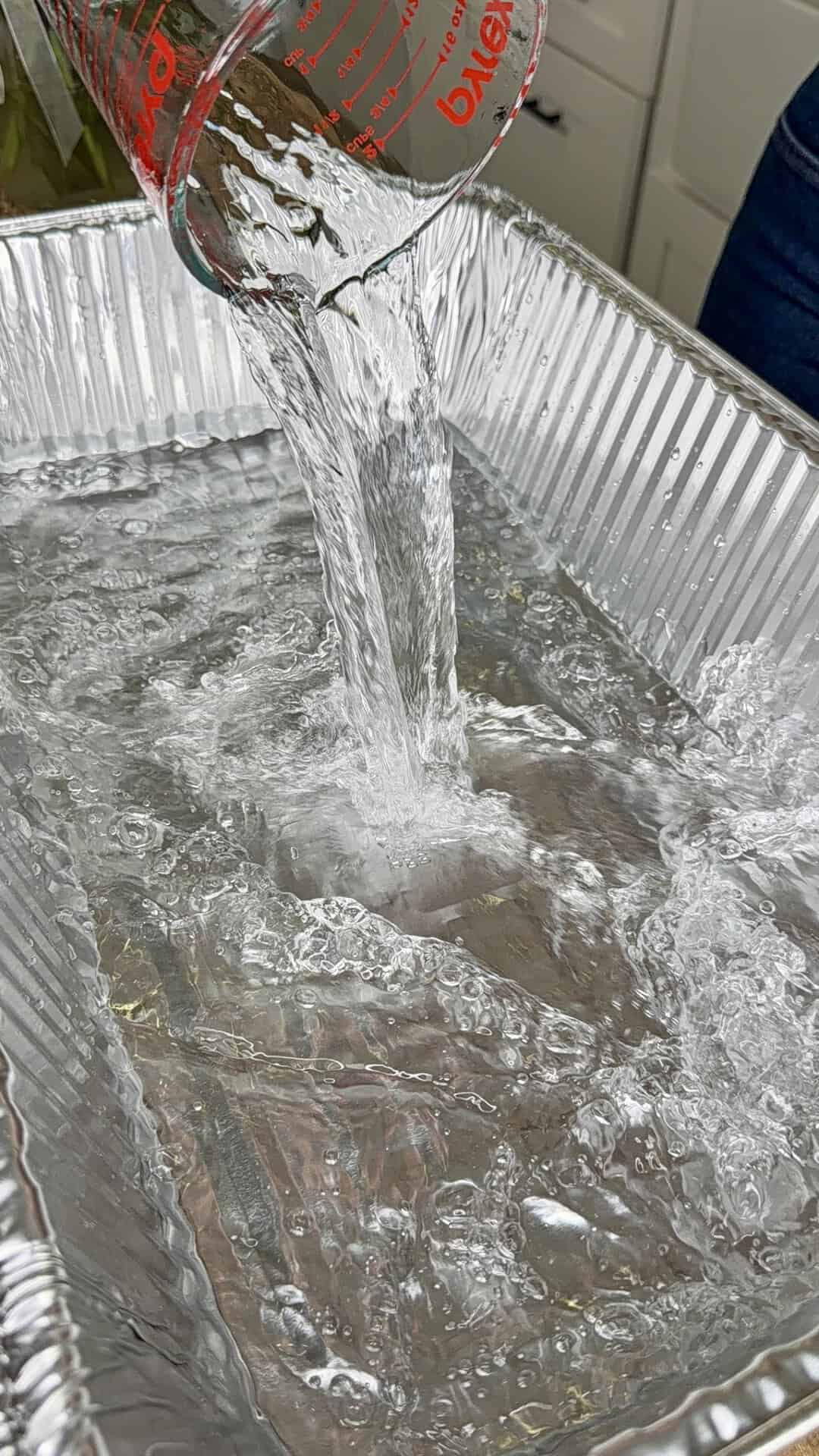 A glass measuring cup pours water into a large, rectangular aluminum tray, causing splashes and ripples across the surface.