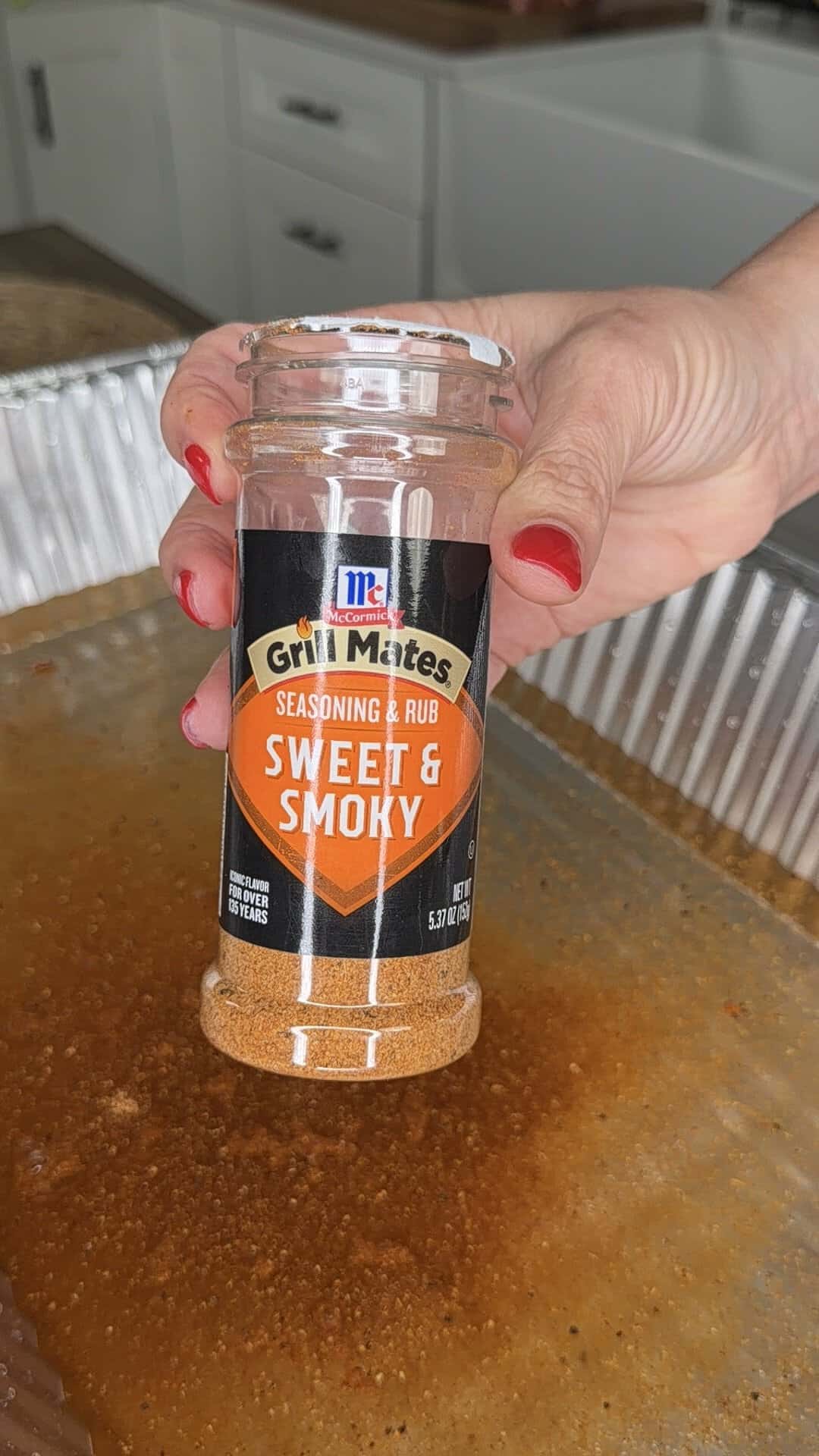 A hand with red painted nails holds a container of McCormick Grill Mates Sweet & Smoky Seasoning & Rub over a metal tray filled with a liquid marinade in a kitchen setting.