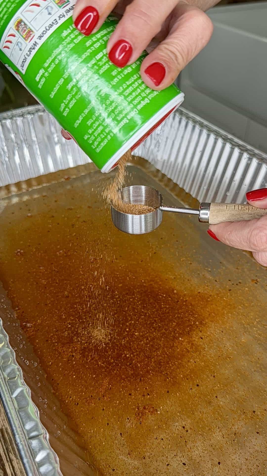 A person with red nail polish pours seasoning from a green can into a metal measuring spoon over a large, shallow aluminum tray containing a brown liquid.